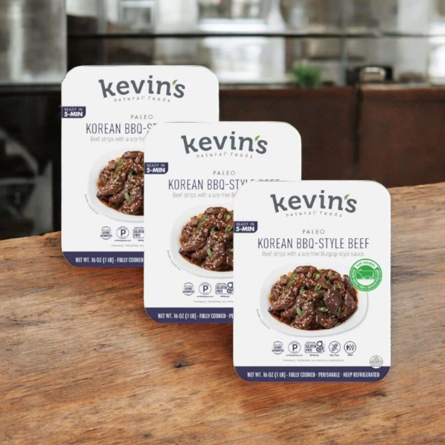 Three packs of Kevin's Paleo Korean BBQ-Style Beef (3 Count) from Kevin's are shown on a wooden surface, with a blurred kitchen in the background. This gluten-free, protein-packed product is perfect for healthy eating.