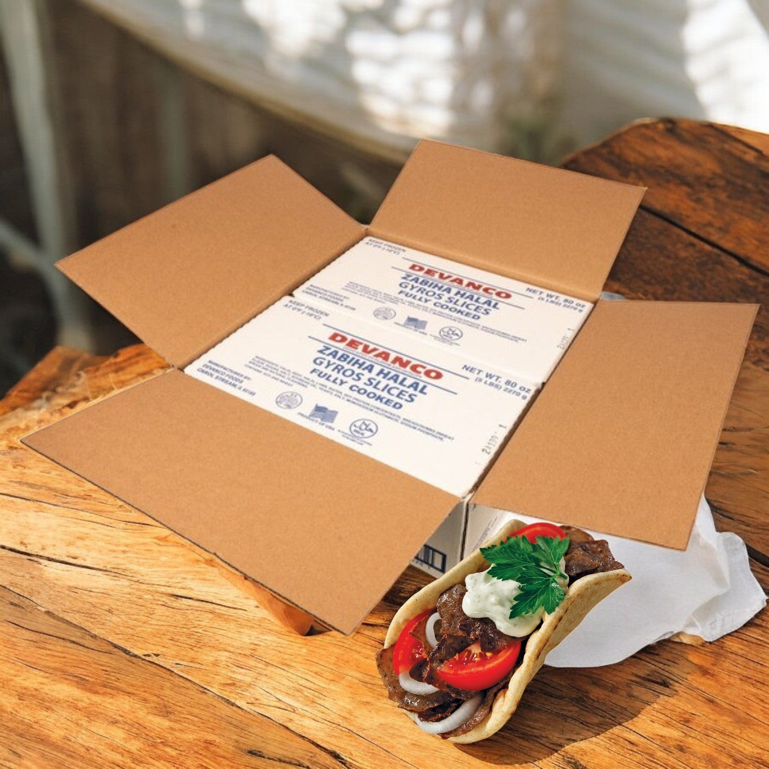 An open cardboard box with two packages of Devanco Foods Halal Gyro Slices (5 lb) sits on a wooden table next to a gyro sandwich filled with beef, lamb, fresh vegetables, and Mediterranean spices.