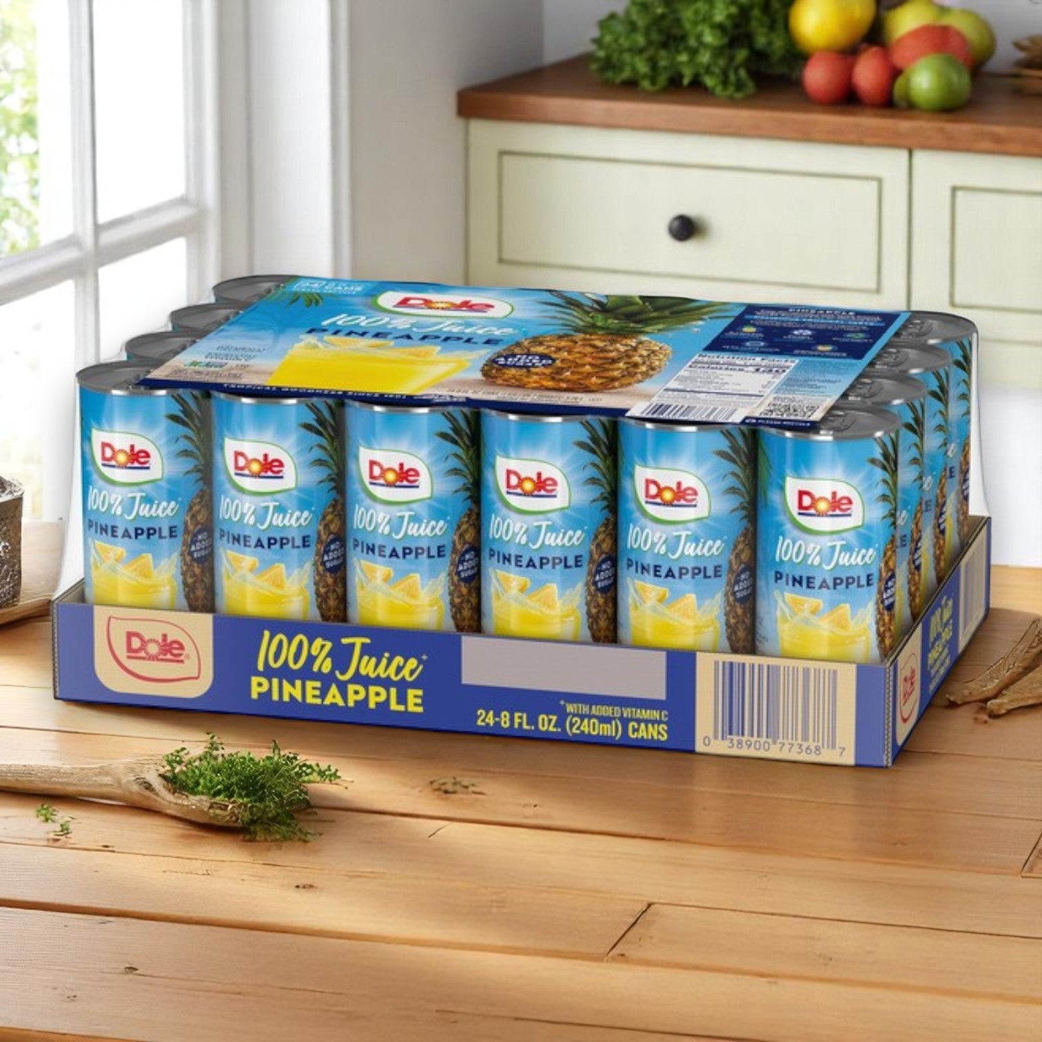 A pack of 24 Dole 100% Pineapple Juice – 8 fl oz cans is displayed on a kitchen counter with fresh fruits and vegetables in the background.