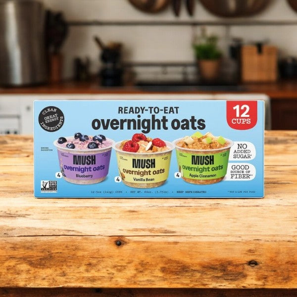 A 12-count box of Mush Overnight Oats Variety Pack (5 oz each) by Mush, featuring blueberry, vanilla bean, and apple cinnamon flavors, is displayed on a wooden kitchen counter.