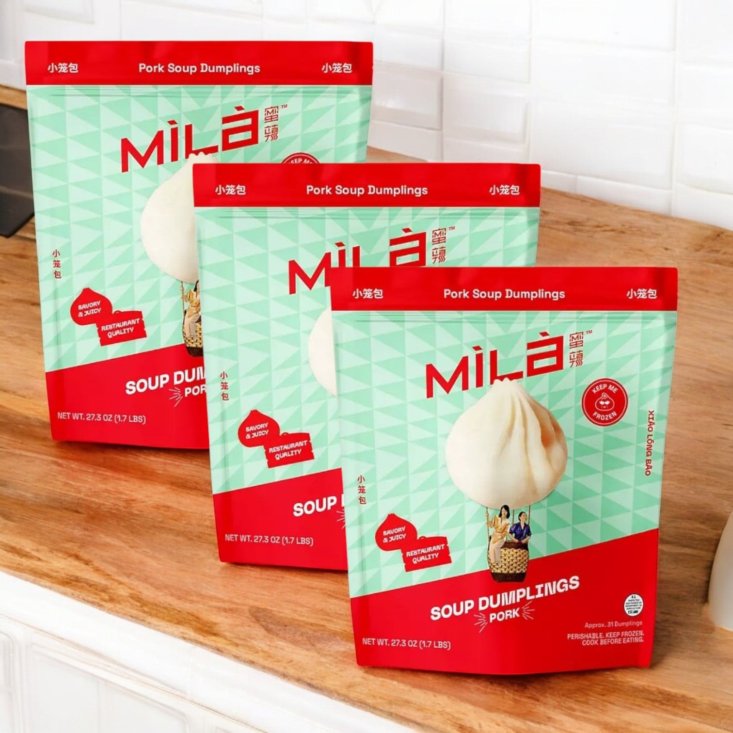 Three Mila Pork Soup Dumplings packages, each with 31 frozen dumplings, sit invitingly on a wooden countertop in the white-tiled kitchen, offering a delicious xiao long bao experience. Perfect for quick meals, they promise authentic flavor with every bite.