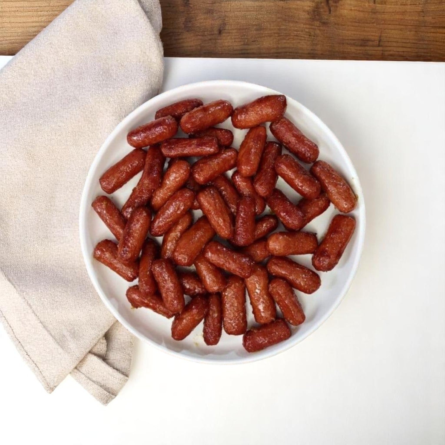 A white bowl holds Nathan's Famous Cocktail Franks – 1200 Count, 1 Case, on a white surface beside a folded beige napkin and a wooden tabletop.