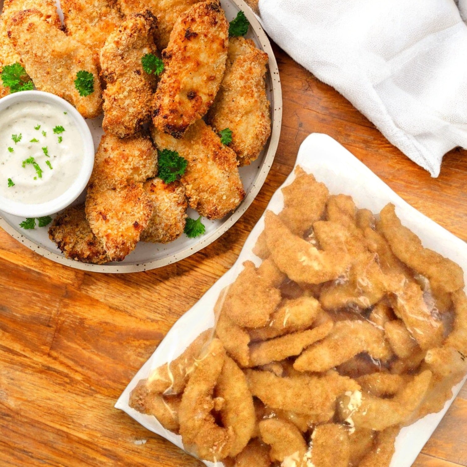 A plate of cooked Sysco Classic Halal Gluten-Free Breaded Chicken Tenders with dipping sauce sits next to a sealed bag of uncooked Sysco Halal Chicken Tenders (2/5 lb) on a wooden table.