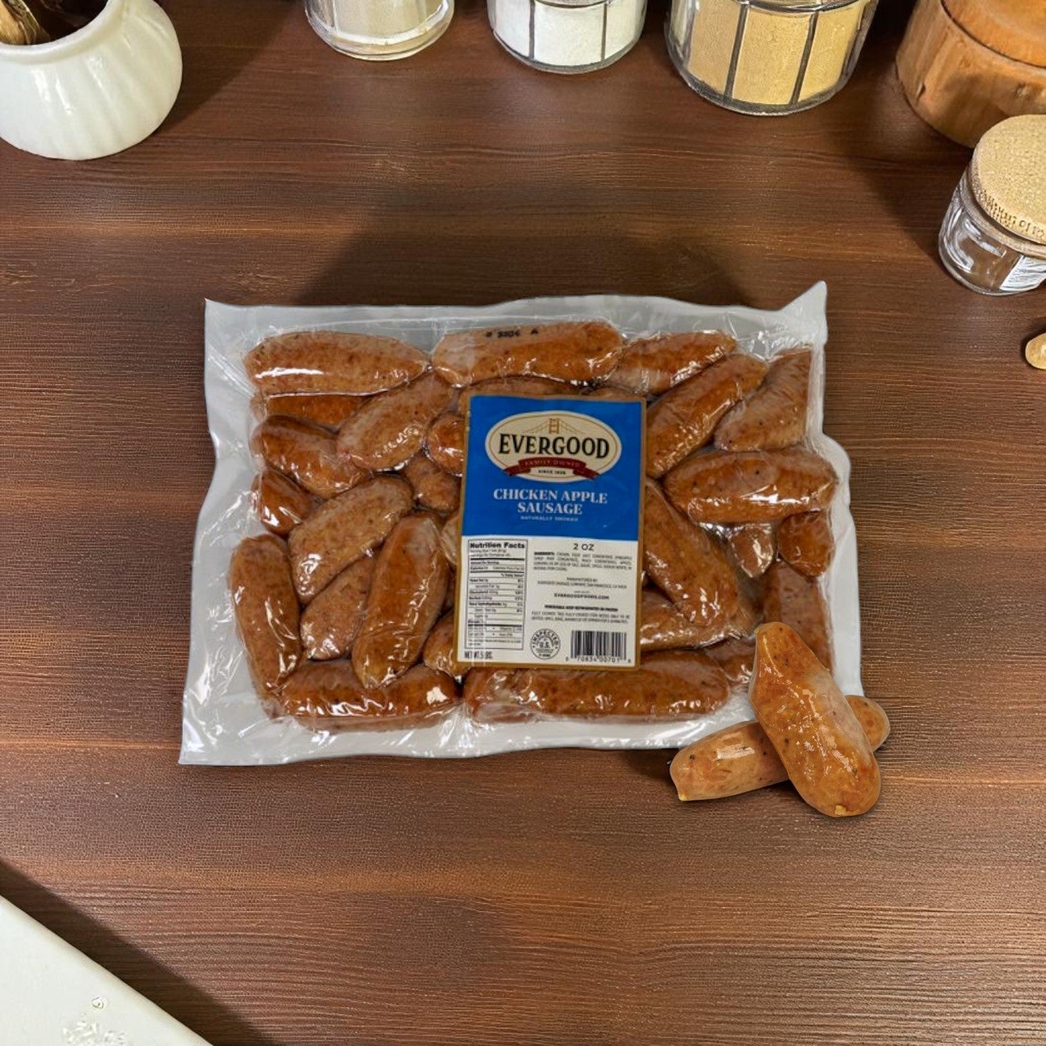 A sealed pack of Evergood Chicken Apple Sausage Links (Frozen, 2 oz links, 2 packs of 5 lbs) rests on a dark wooden countertop with two sausages outside the package.