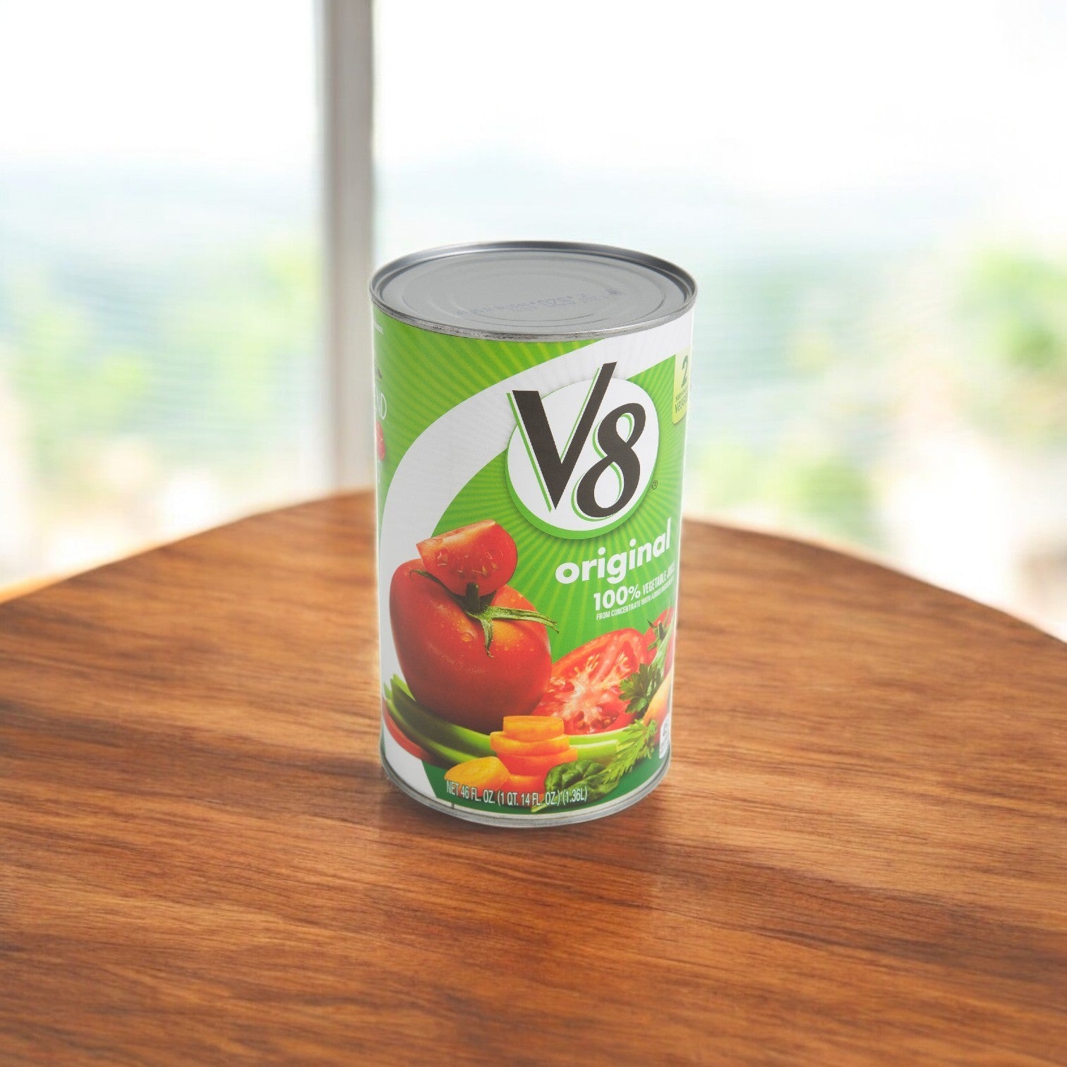 A V8 Vegetable Juice can from a 46 oz., 1 Case, 12 Pack sits on a wooden table against a blurred outdoor backdrop, presenting a heart-healthy choice with its 100% vegetable juice.