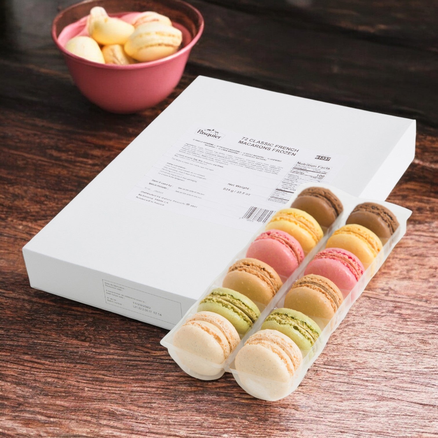 A Pasquier Cookie Mini Assorted Macarons box, 1 Case with 72 pieces, adorns a wooden table, complemented by a pink bowl filled with gourmet macarons in the background.