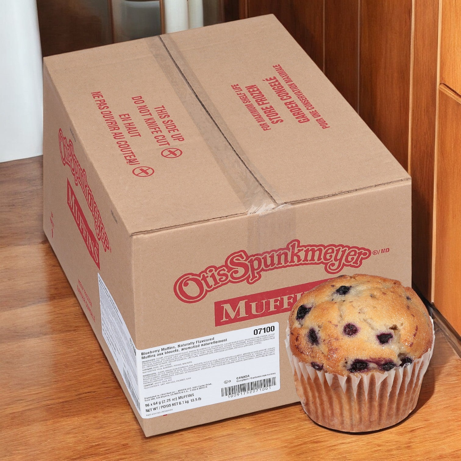 A large cardboard box labeled "Otis Spunkmeyer Muffin Blueberry Wild, 1 Case, 96 Total Count—Free Shipping!" is on a wooden floor with a Wild Blueberry Muffin in front, ideal for a breakfast snack.