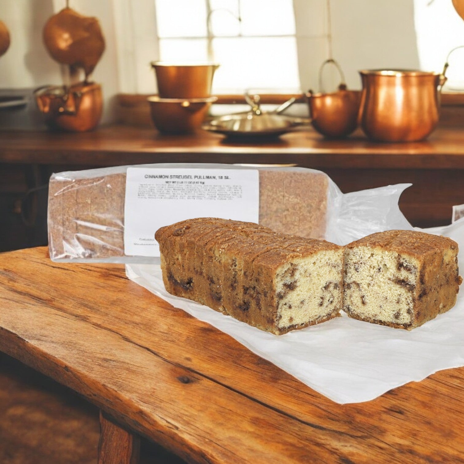 The Cake Loaf Cinnamon Streusel Slices from Sysco, resting on parchment paper, fill the kitchen with their aroma amidst gleaming copper pots. Enjoy these cozy dessert slices on a wooden table. Each pack includes 2 loafs of 12 inches, totaling 36 pieces!.