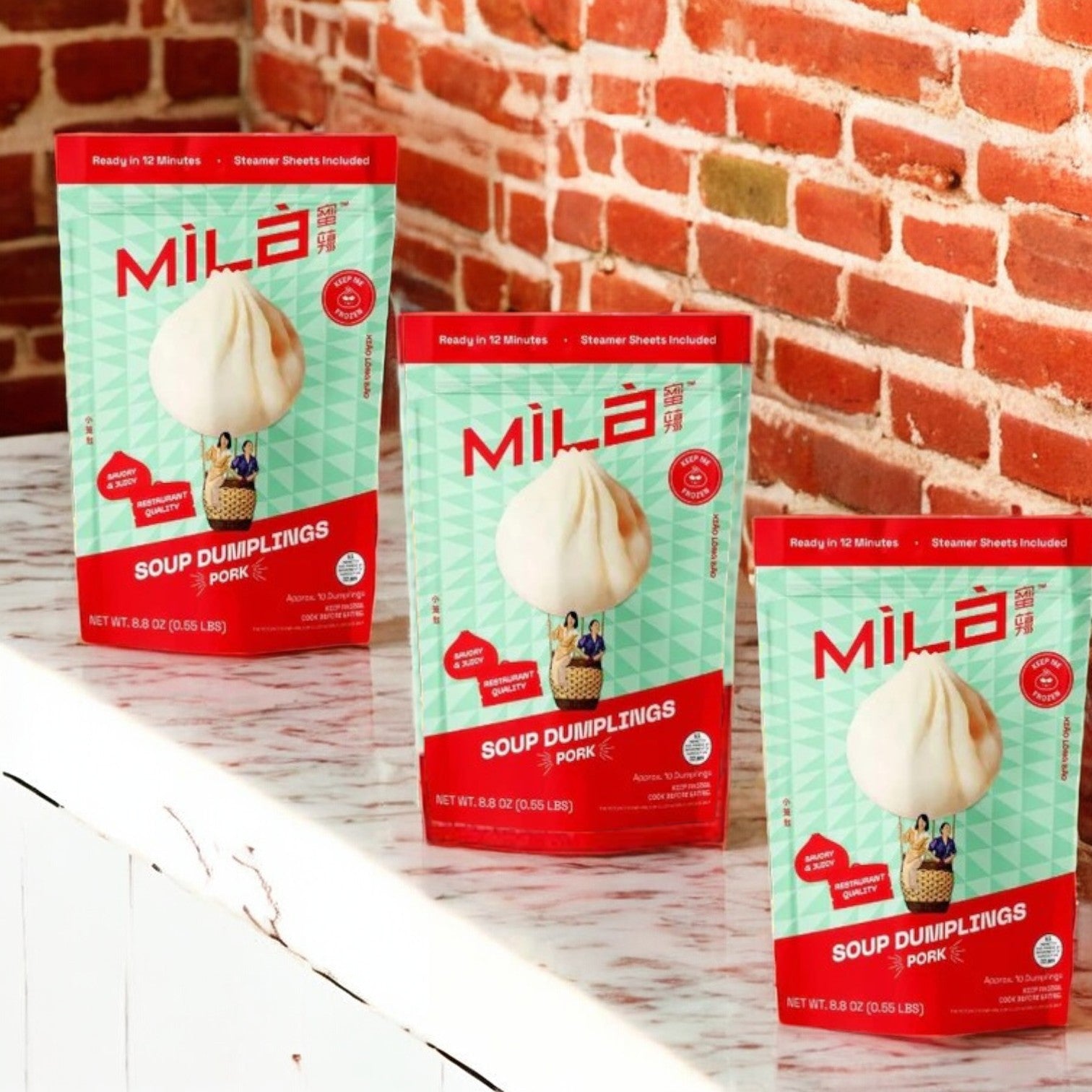 Three packages of MiLa Frozen Soup Dumplings - Pork - 3 Count are displayed on a white marble surface with a red brick wall in the background.
