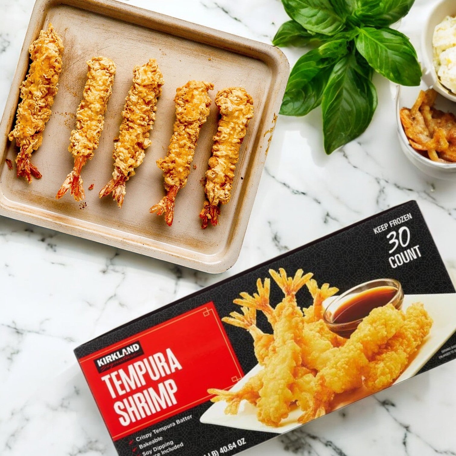 Five Kirkland Signature Tempura Shrimp sit on a tray next to a box of Kirkland Signature Tempura Shrimp, 2.35 lbs, 30-count—garnished with basil and served with dipping sauce on a marble countertop.