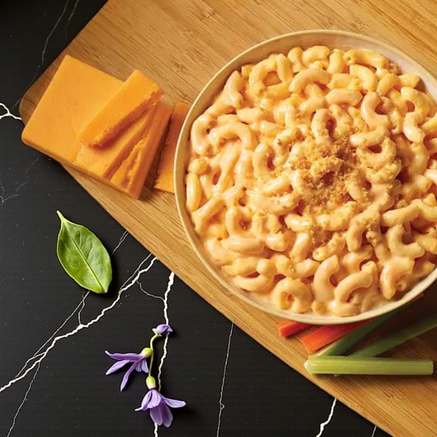 TJ Ribs Mac & Cheese – 6 oz by TJ Ribs features creamy mac and cheese with breadcrumb topping, served on a wooden board alongside cheddar slices, celery sticks, a green leaf, and a purple flower—ideal as an individual side or comfort food.