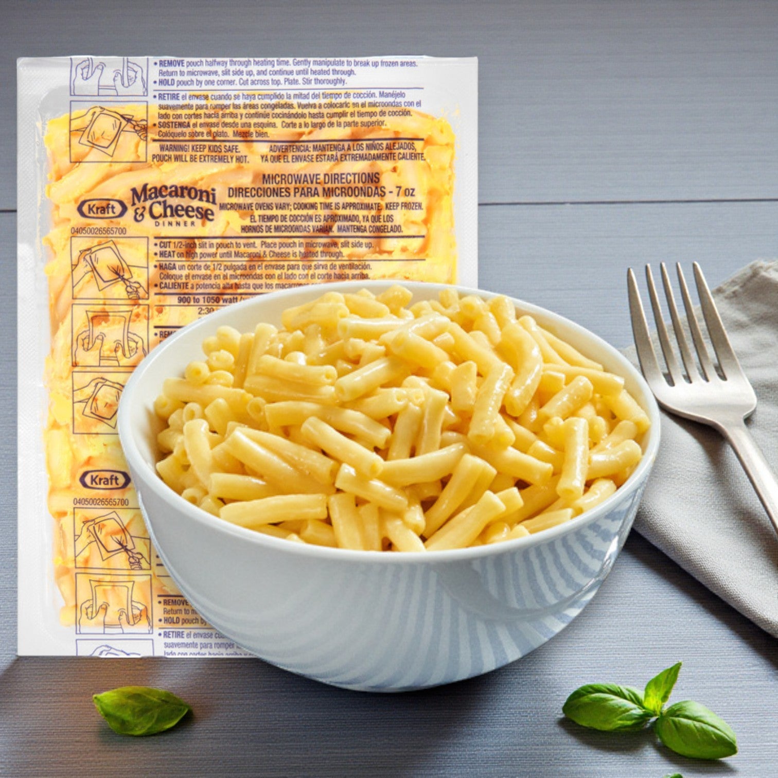 A bowl of Kraft Frozen Entree Macaroni & Cheese – 36 Packs (7 oz per pack) by Kraft sits on a table with a fork, napkin, and basil leaves; the empty packaging and instructions are visible in the background.