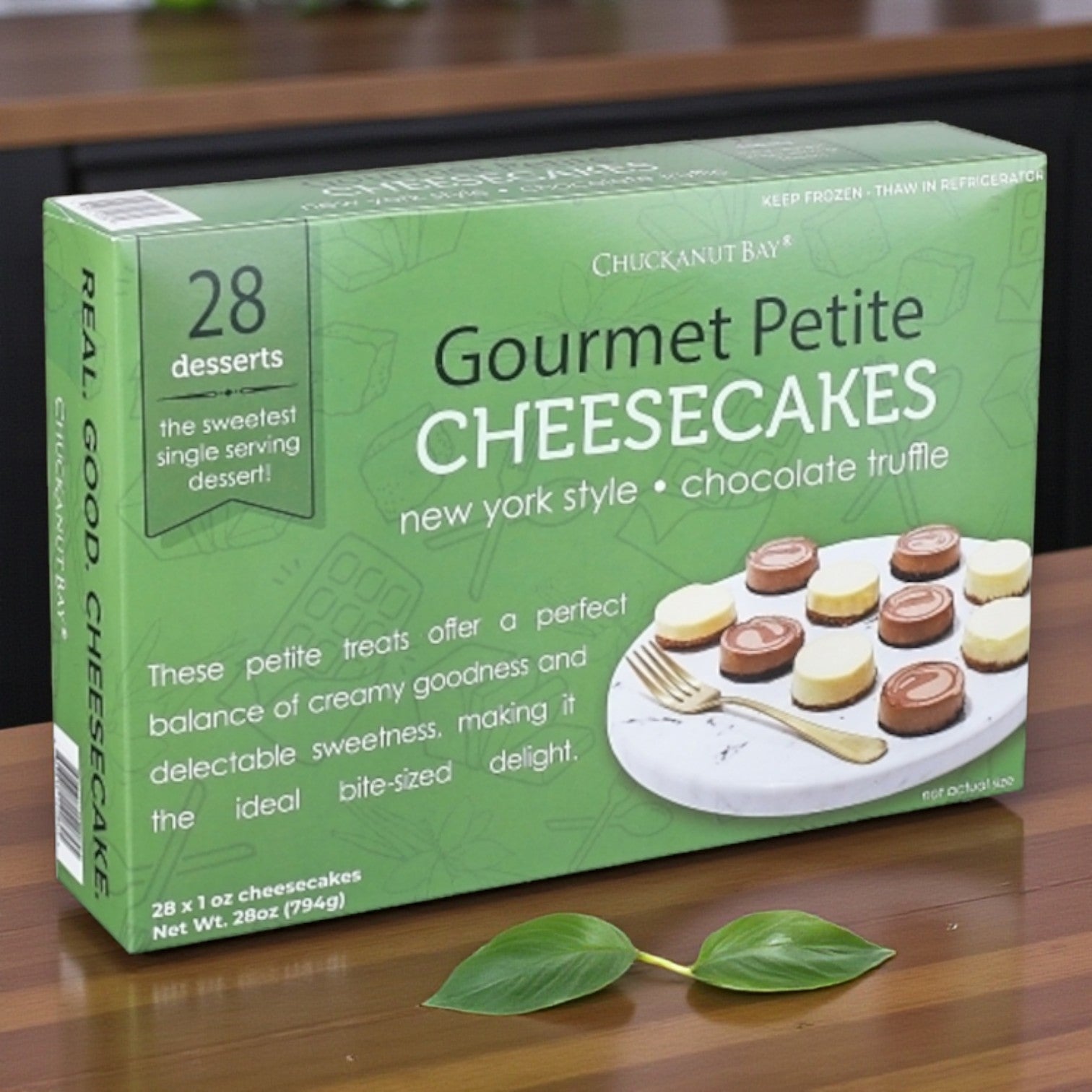 A box of Chuckanut Bay Assorted Mini Cheesecakes – 28 Count sits on a counter, with two green leaves in front.