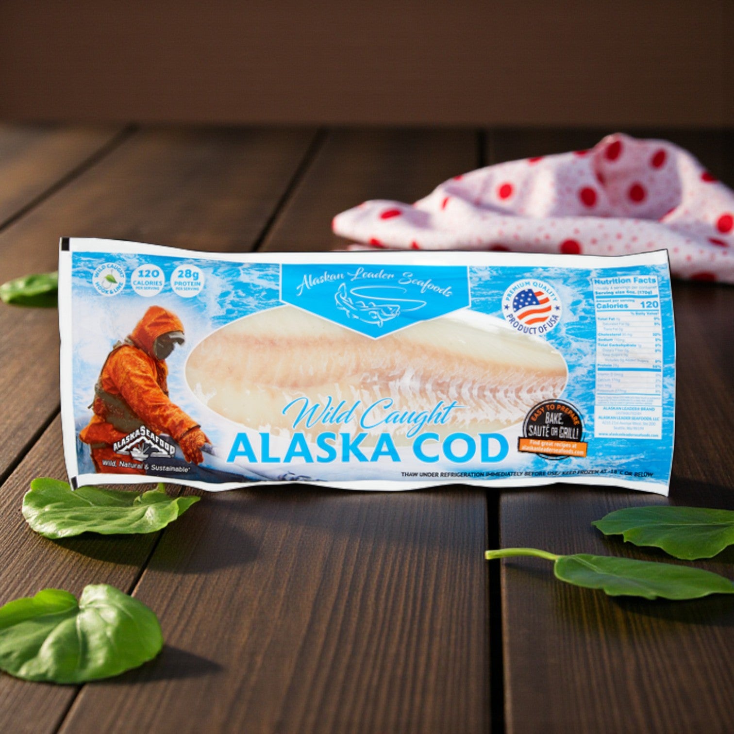 A 2lb package of Alaskan Leader Seafoods Alaska Cod Fillet, Wild Caught, rests on a wooden surface with spinach leaves and a red polka dot cloth nearby.