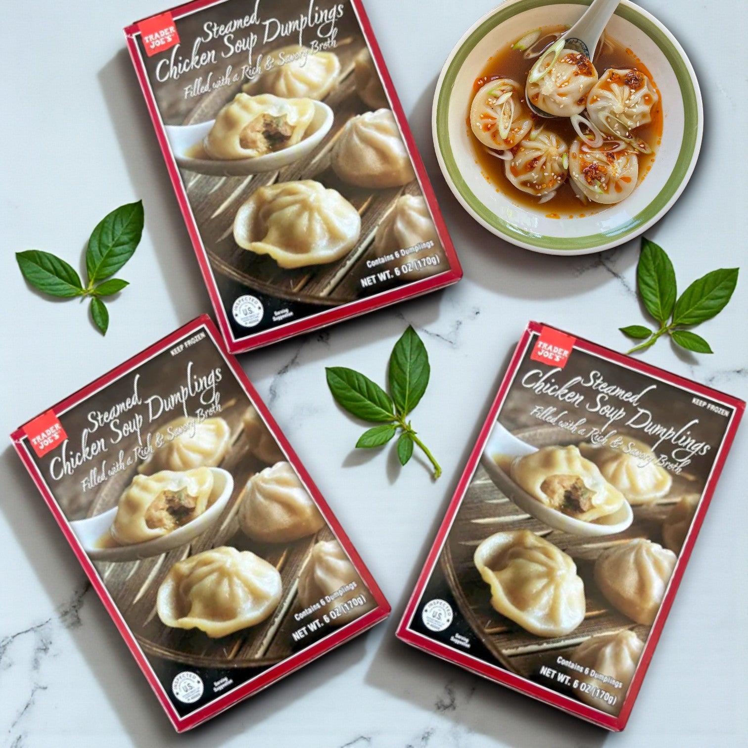 Three packs of Trader Joes Steamed Chicken Soup Dumplings – 6 oz are displayed on a surface, alongside fresh basil leaves, a bowl of chicken soup dumplings in broth, and a spoon.