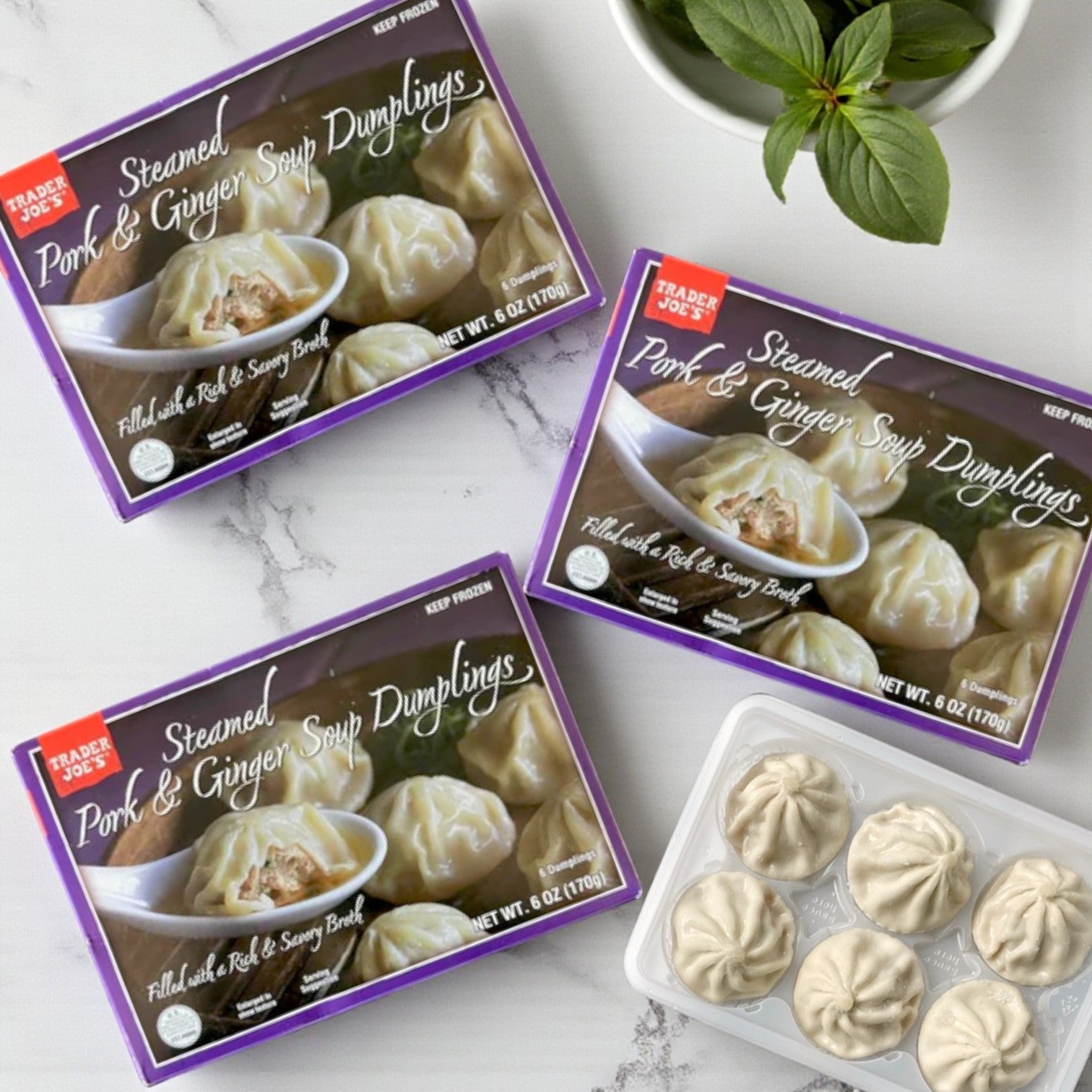 Three boxes of Easy Lunches Trader Joe’s Steamed Pork & Ginger Soup Dumplings – 6 oz (3 Pack) are shown on a white surface, with one open to reveal six dumplings in a tray and a sprig of basil beside it.