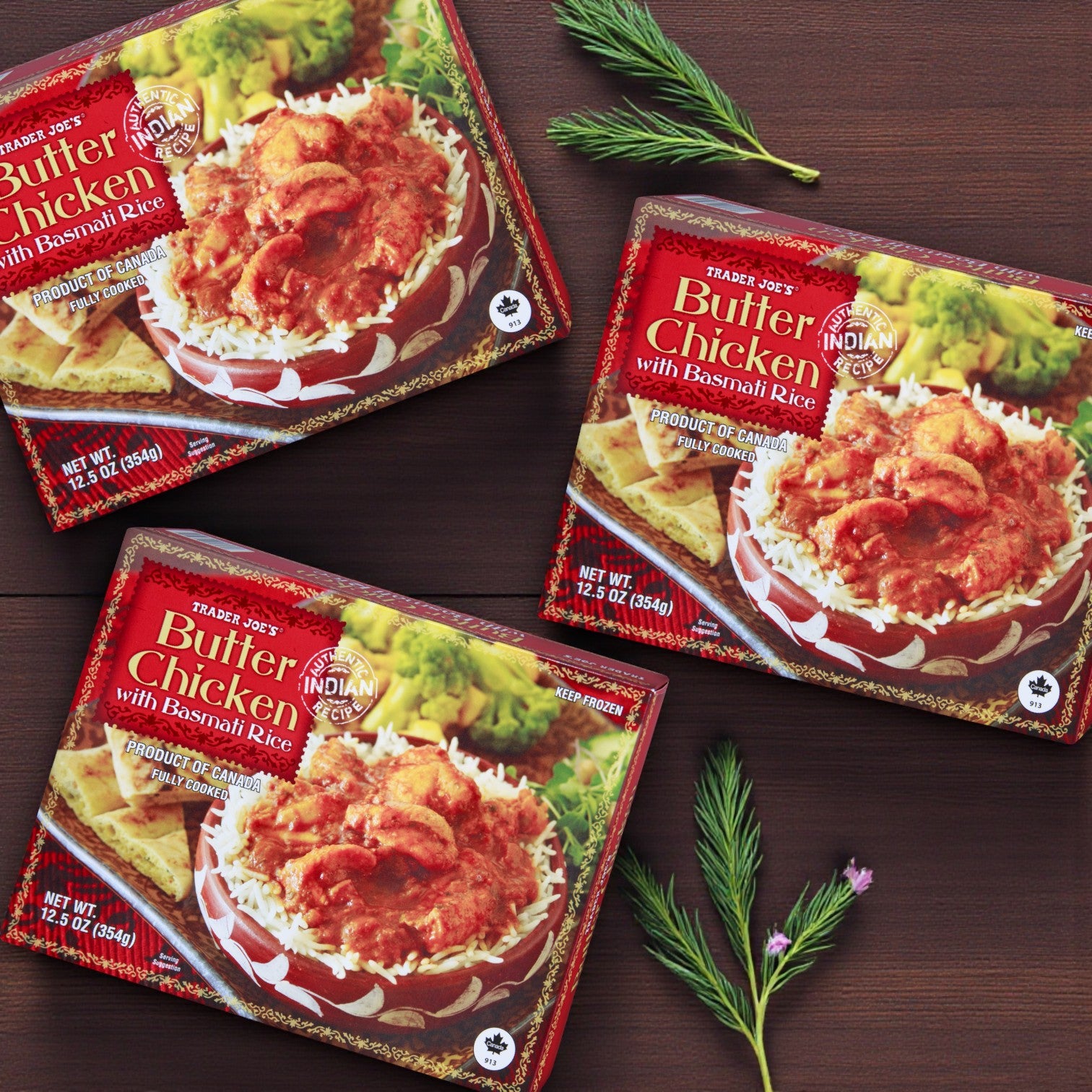 Three packs of Easy Lunches Trader Joe’s Butter Chicken with Basmati Rice – 12.5 oz are displayed on a wooden surface decorated with green sprigs and a small purple flower.
