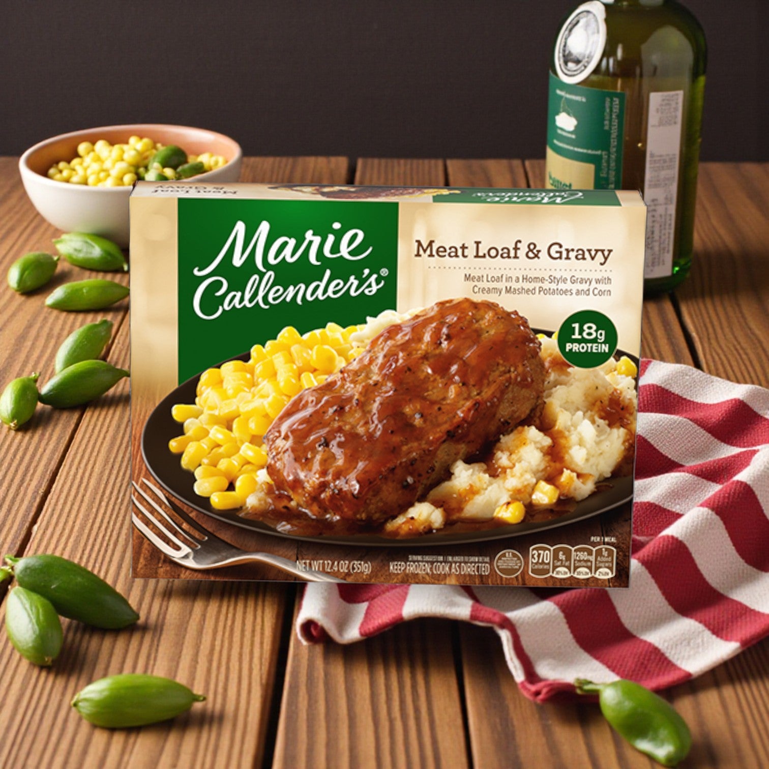 A Marie Callender’s Comfort Classics Entrée Combo – 5 Delicious Meals sits on a wooden table with a red checkered napkin, corn, green peppers, and a bottle in the background.