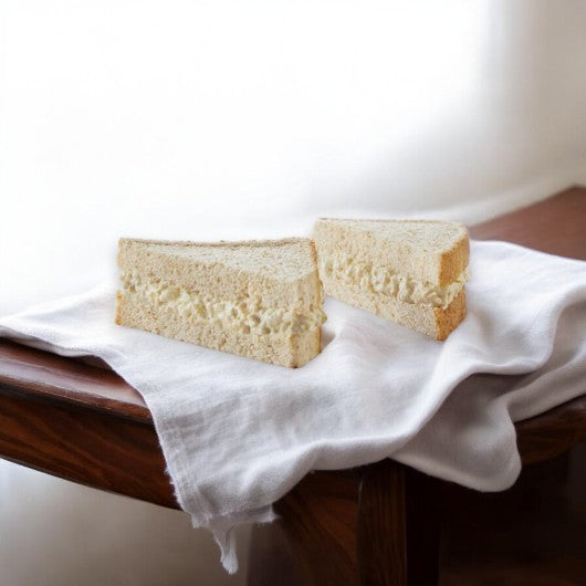 Two E.A. Sween Albacore Tuna Salad Sandwiches from a case of 8 are shown on a white cloth atop a wooden table, with soft natural light highlighting their freshness. Ships frozen; once thawed, enjoy up to 30 days shelf life in the cooler!.