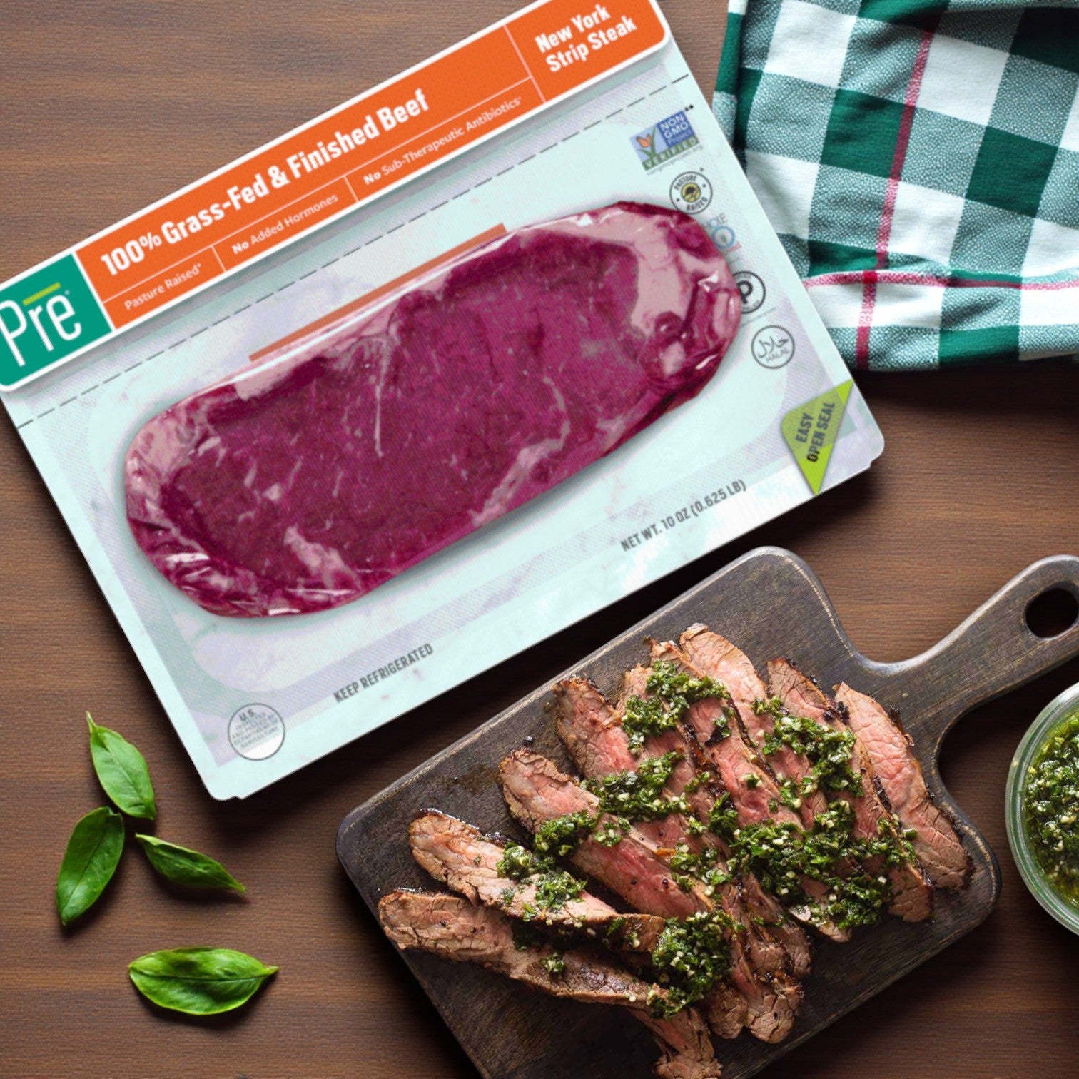 A package of Pre Brands 100% Grass Fed New York Strip Steak – 10 oz (Fresh Beef) is next to a cutting board with cooked, sliced steak topped with green sauce, beside a checkered napkin and basil leaves.