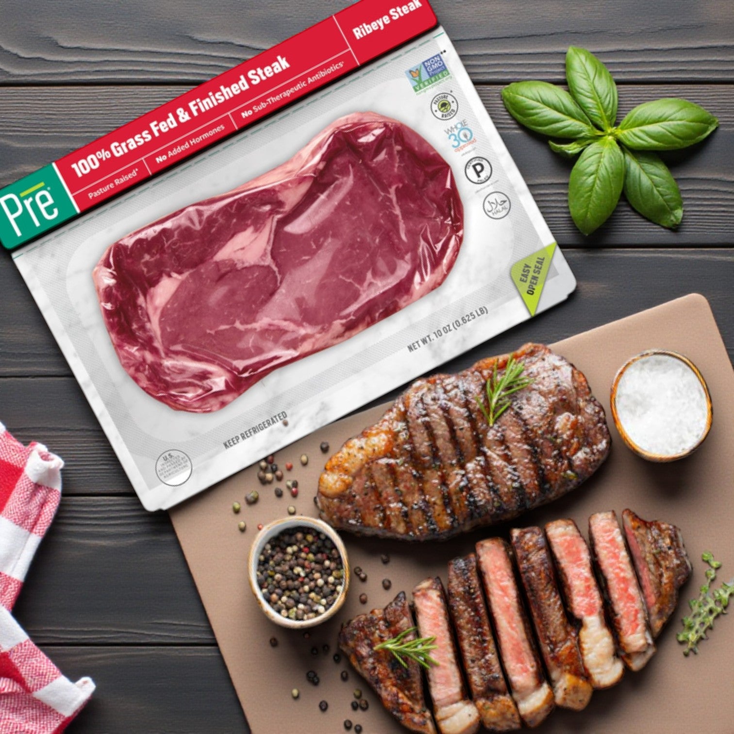 A 10 oz. PRE Brands Ribeye Steak—100% Grass Fed Fresh Beef—sits by a cutting board with sliced steak, basil, salt, and peppercorns, ideal for those who savor premium quality beef from Pre Brands.