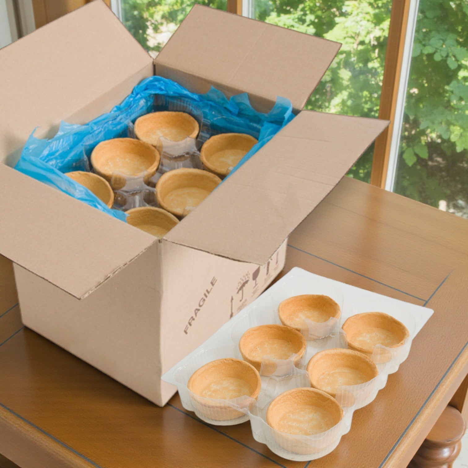 A case of Pidy Inc. Pastries’ 4.5" baked quiche shells (42 count) in a plastic tray sits on a wooden table beside another tray of ready-to-fill Pidy pastry shells.