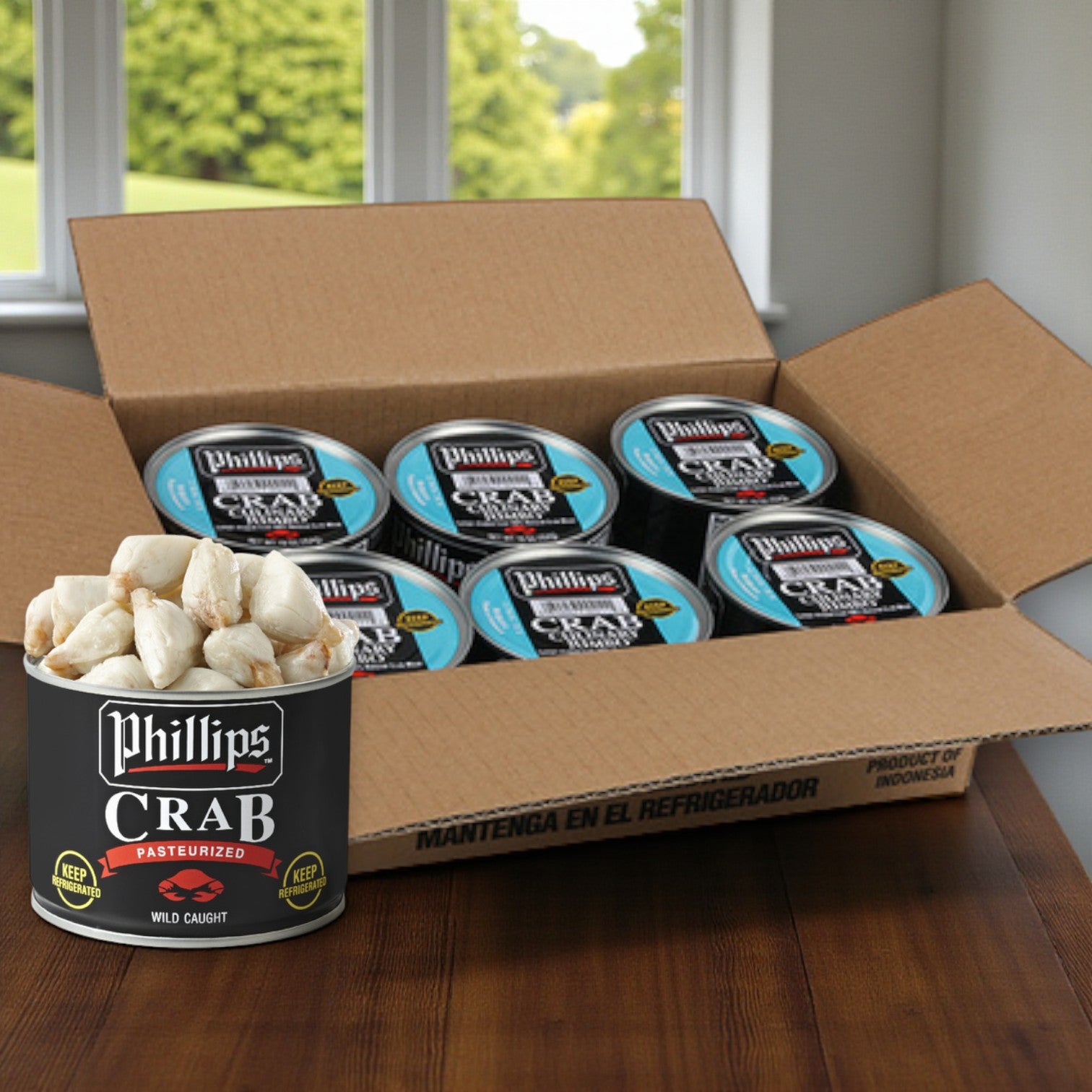 A case of six 1 lb cans of Phillips Foods Pasteurized Jumbo Lump Crab Meat sits open on a wooden table by a window with a green outdoor view.