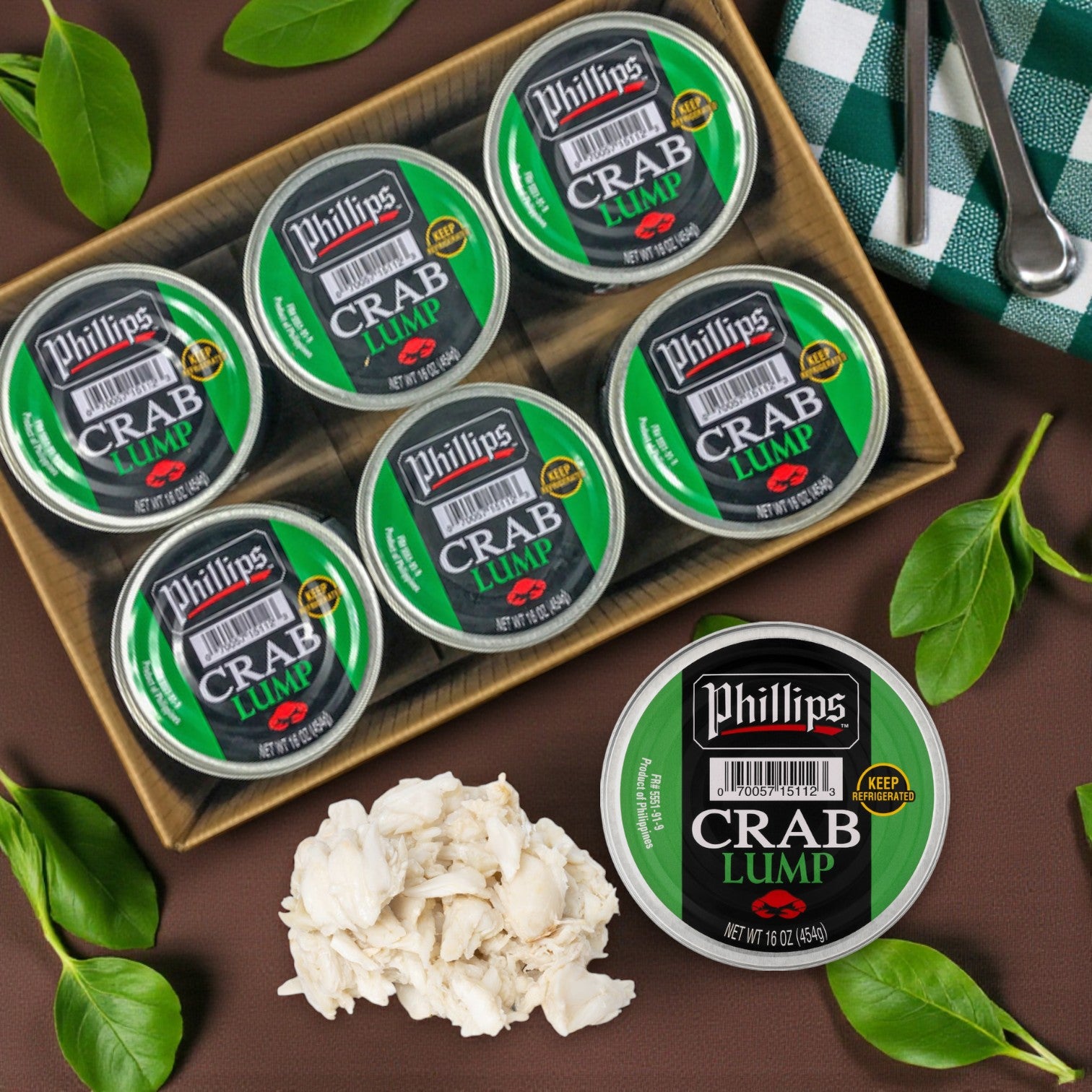 Six 1 lb cans of Phillips Foods Pasteurized Lump Crab Meat are arranged in a tray, with one open to show the tender lump crab. Green leaves and a checkered cloth add accents on a brown surface. Available in a 6 count case.