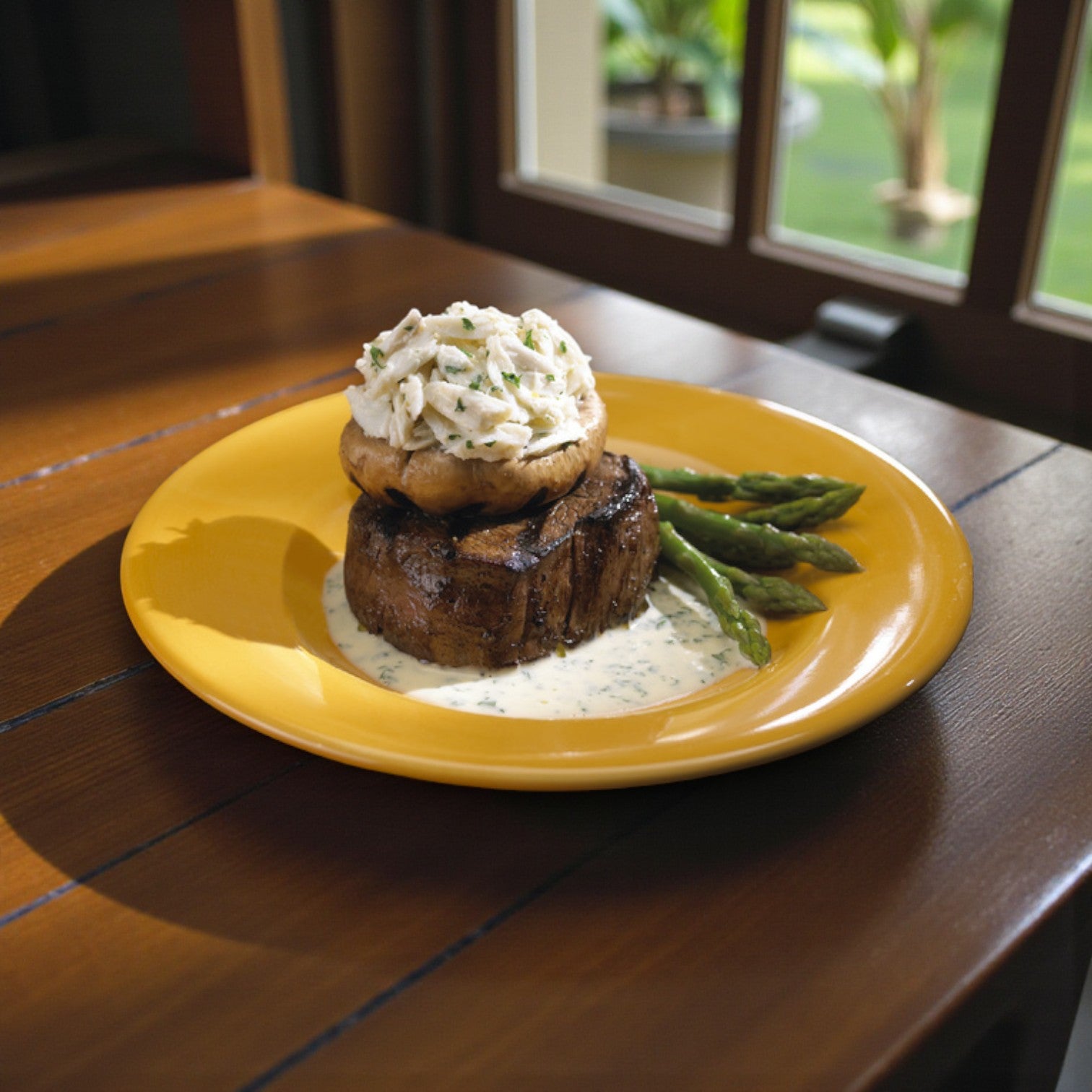 Grilled steak topped with a stuffed mushroom and creamy white sauce made with Phillips Foods Pasteurized Lump Crab Meat, served alongside asparagus on a yellow plate by a sunlit window.