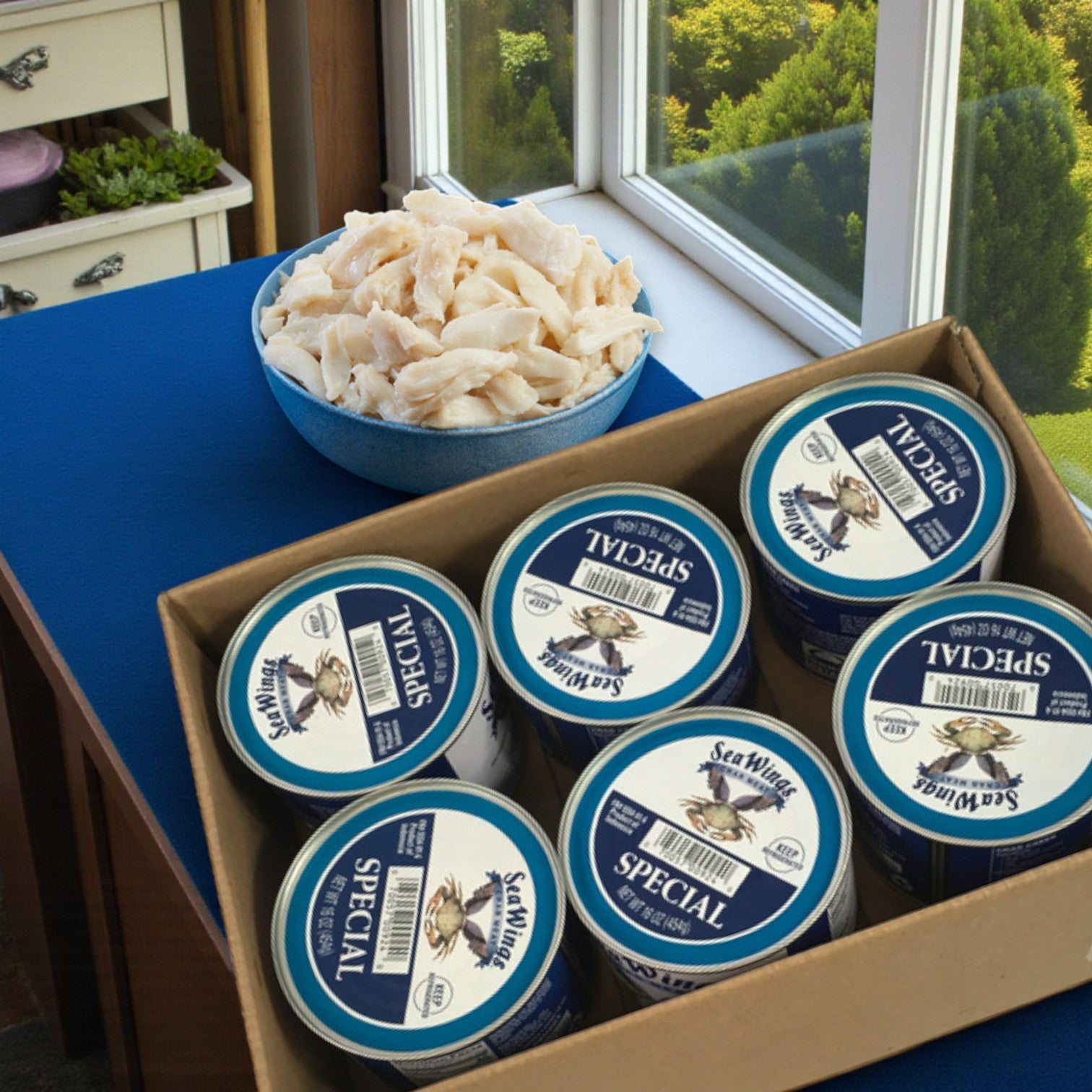 A bowl of Sea Wings Pasteurized Special Crab Meat sits next to a 6-count case of 1 lb cans on a blue table by the window, perfect for seafood stuffing recipes.