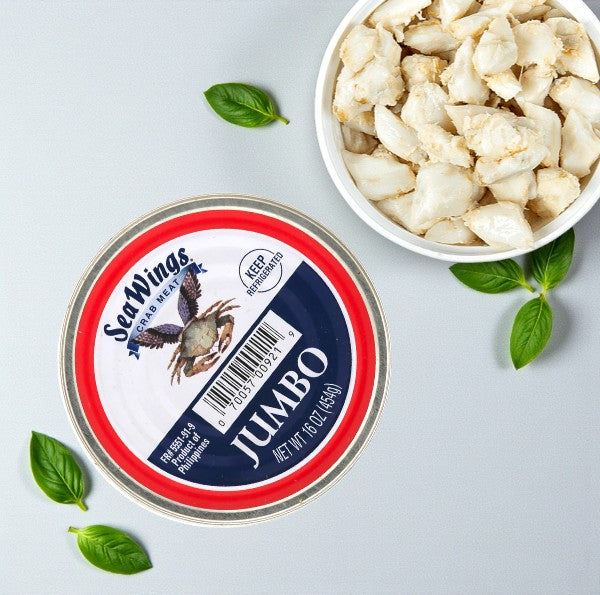 A can of Sea Wings Pasteurized Jumbo Lump Crab Meat (1 lb) beside a bowl of crab meat, with fresh basil leaves scattered on a light surface. Comes in a 6 count case from Sea Wings.