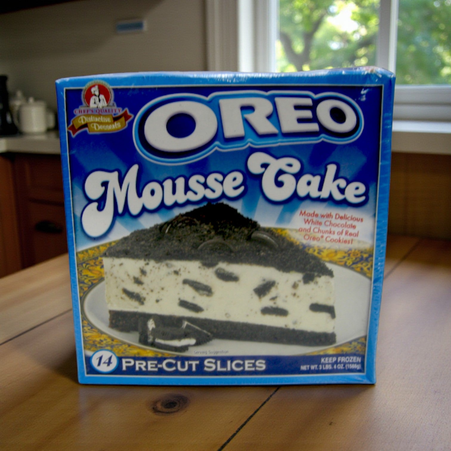 A box of Easy Lunches Frozen Chef’s Quality Oreo Mousse Cake with 14 pre-sliced pieces of rich cookies & cream mousse, displayed on a wooden table in a kitchen.