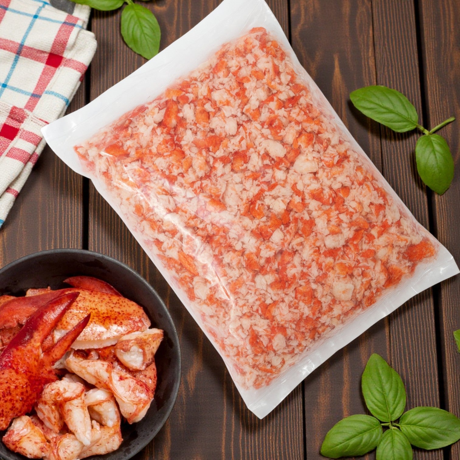 A vacuum-sealed pack of Packer Lobster Meat – Claw & Knuckle (4 lbs) sits on a wooden table next to a bowl of lobster pieces and fresh basil leaves.