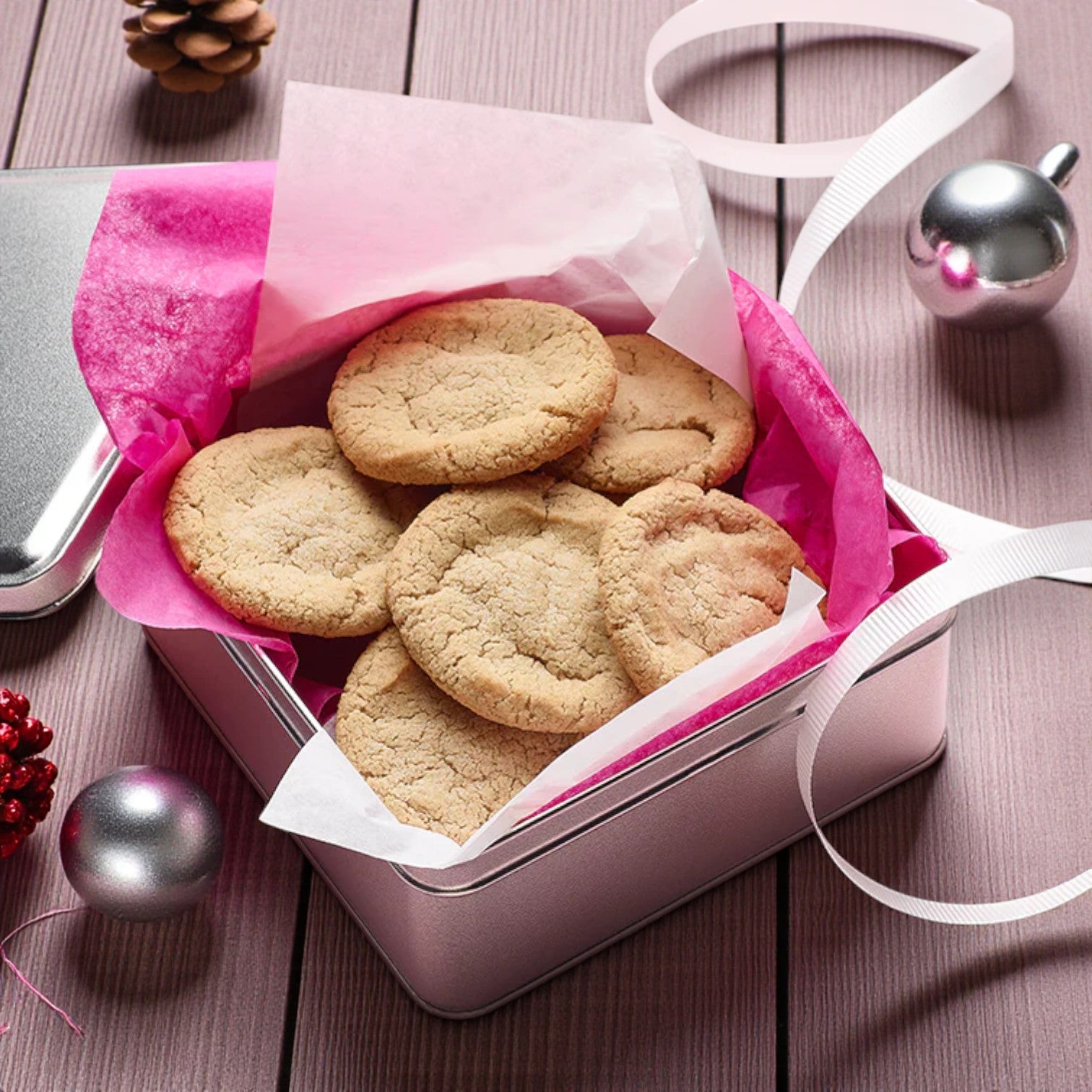 A silver tin lined with pink tissue cradles six sugar cookies baked from Sweet Loren’s Sugar Cookie Dough – Gluten-Free & Vegan 12 oz (3-Pack), by Sweet Loren's, surrounded by festive ornaments, ribbon, and a pine cone on a wooden table.