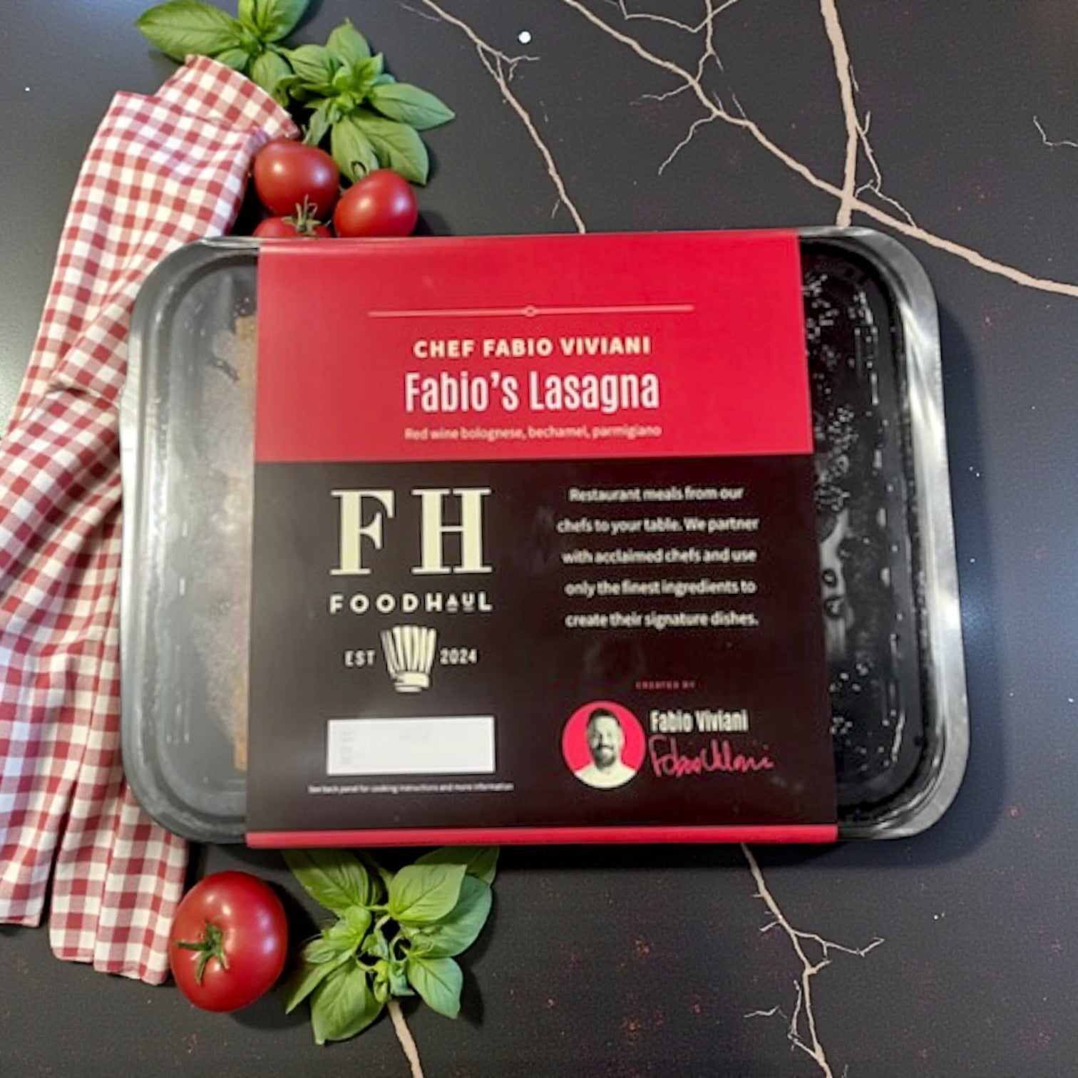 A FoodHaul Frozen Meal – Fabio’s Lasagna by Fabio Viviani rests on a counter with tomatoes, basil, and a checkered napkin. The packaging features quality ingredients, Italian comfort food vibes, and Chef Viviani’s photo.