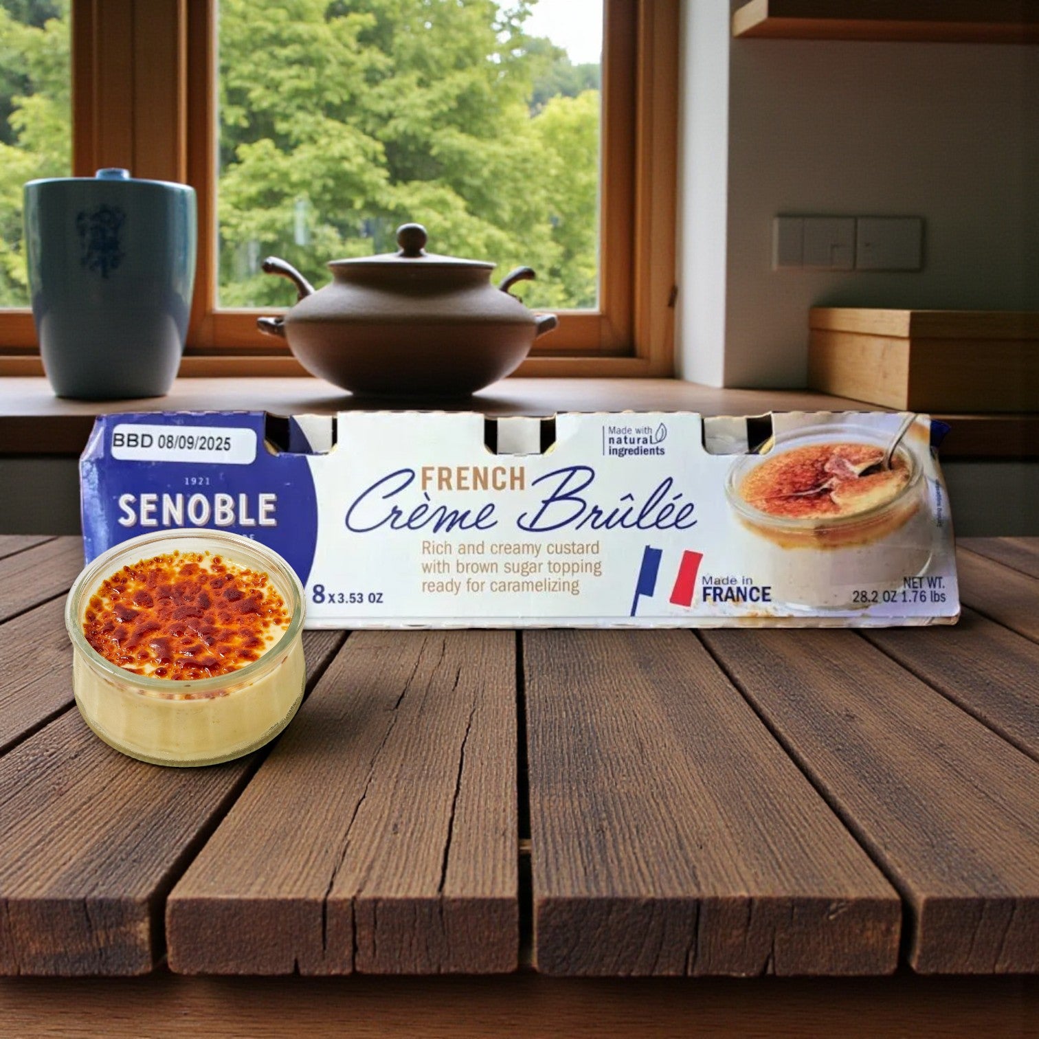 A package of Senoble French Crème Brûlée, 3.75 oz, 8-Count, sits on a wooden table alongside a glass dish of vanilla custard with caramelized sugar topping in a kitchen with a window view.