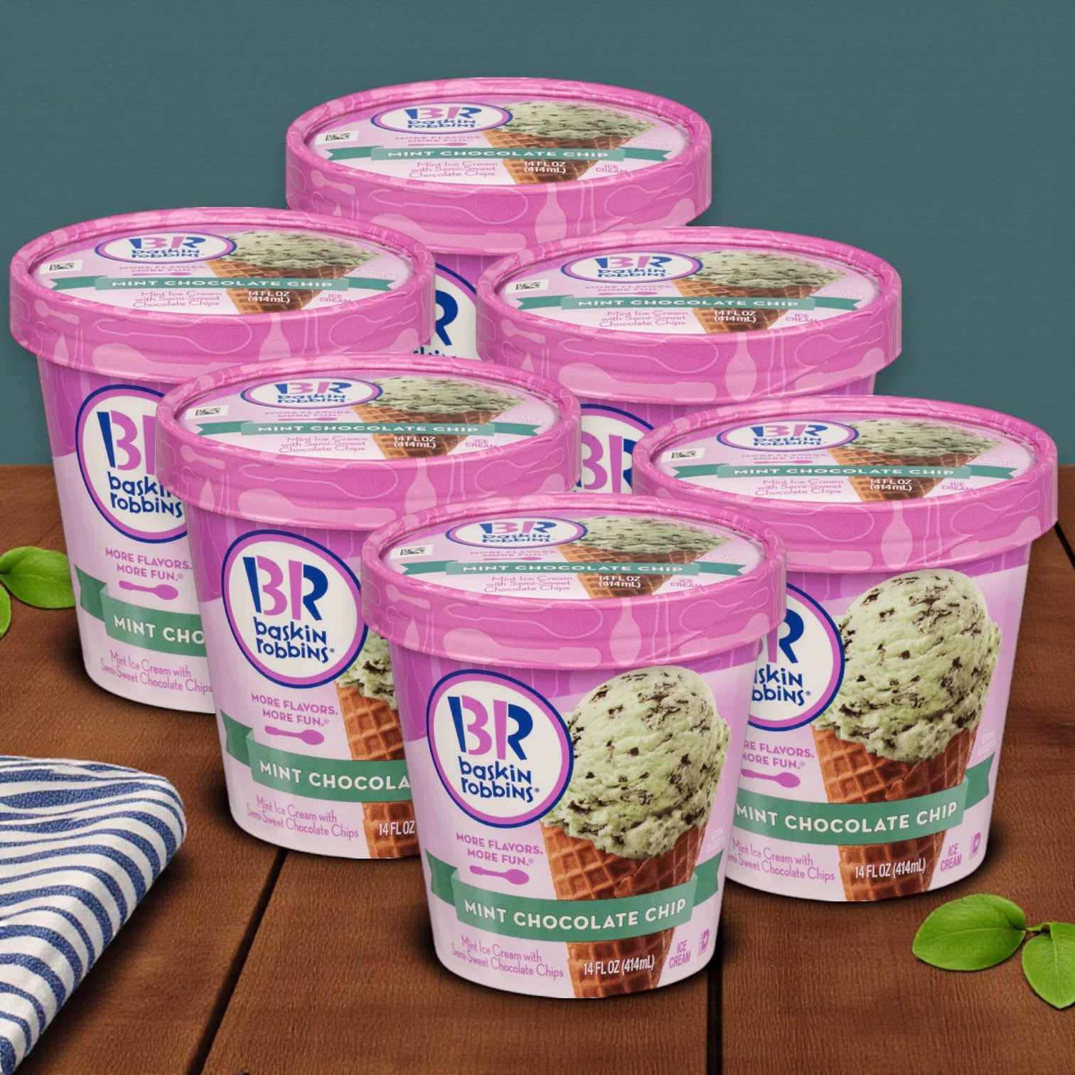 Six 14 oz. pints of Baskin Robbins Ice Cream Mint Chocolate Chip are displayed on a wooden surface with a blue striped cloth and green leaves, offering a fresh and inviting showcase of this premium dessert.