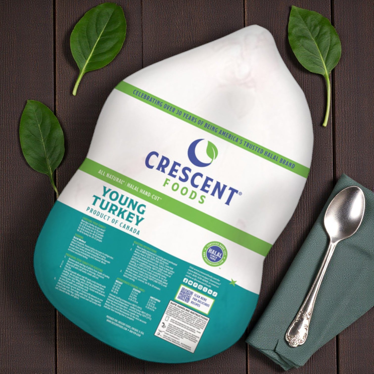 A frozen Crescent Foods All-Natural Halal Young Turkey (12-14 lb average) sits on a dark surface with green leaves, a folded napkin, and a spoon nearby.