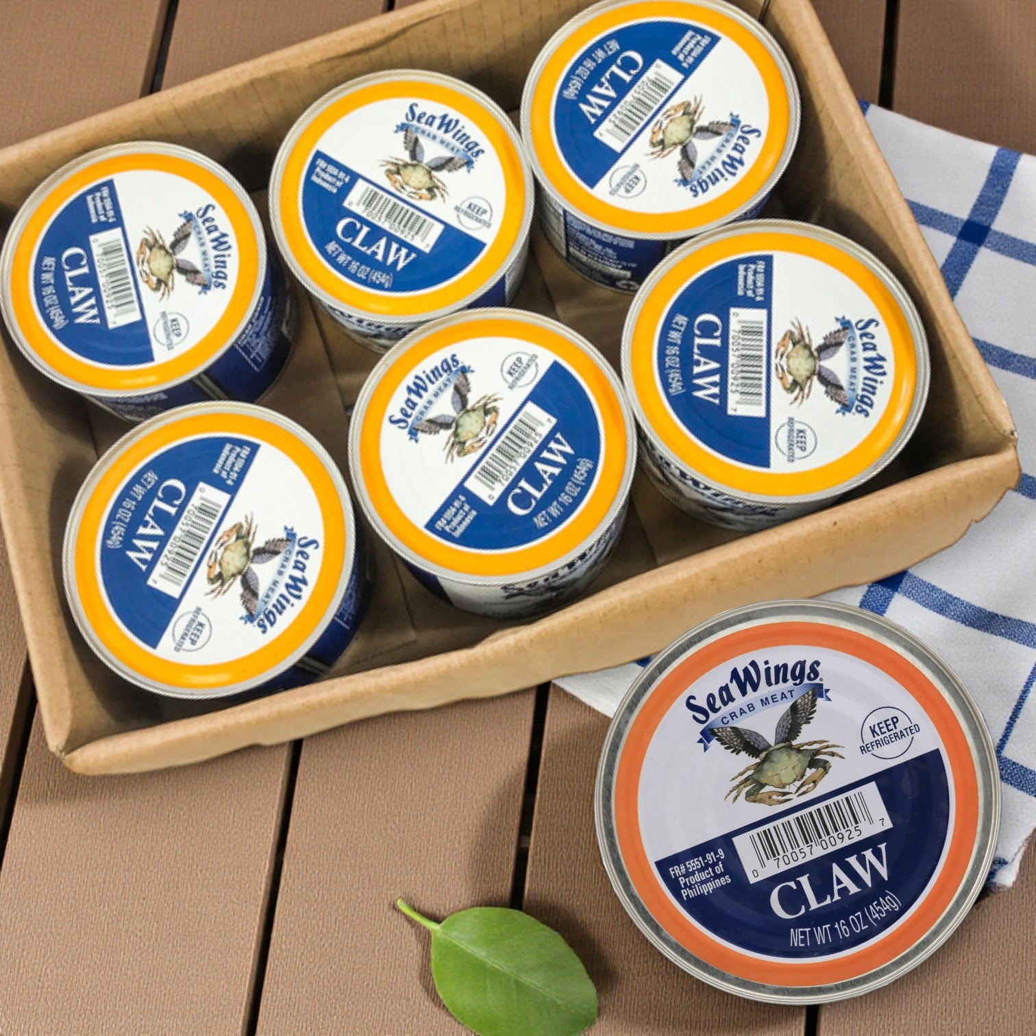 A cardboard tray holds six 1 lb cans of Sea Wings Pasteurized Claw Crab Meat, next to a white and blue cloth and a green leaf.