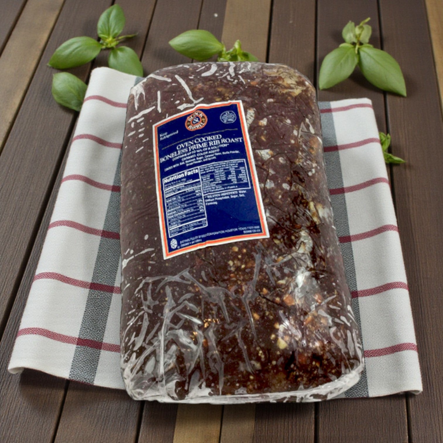 A Sysco Block & Barrel Classic Cooked Boneless Beef Prime Rib (very rare, 15%), 11–14 lbs per piece, is displayed on a white and red striped towel with green basil leaves atop a wooden surface. Pack: 2 pieces per case.