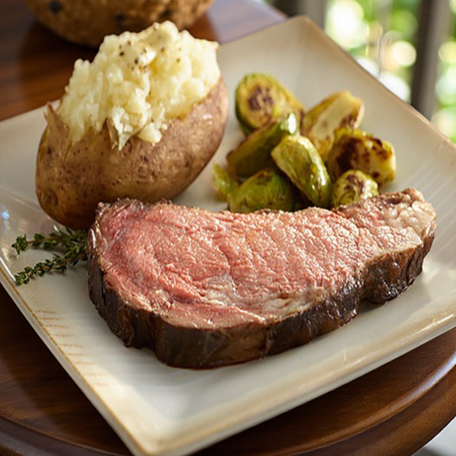 A plate featuring Sysco's Block & Barrel Classic beef prime rib (boneless, very rare), served with a baked potato topped with butter and roasted Brussels sprouts.