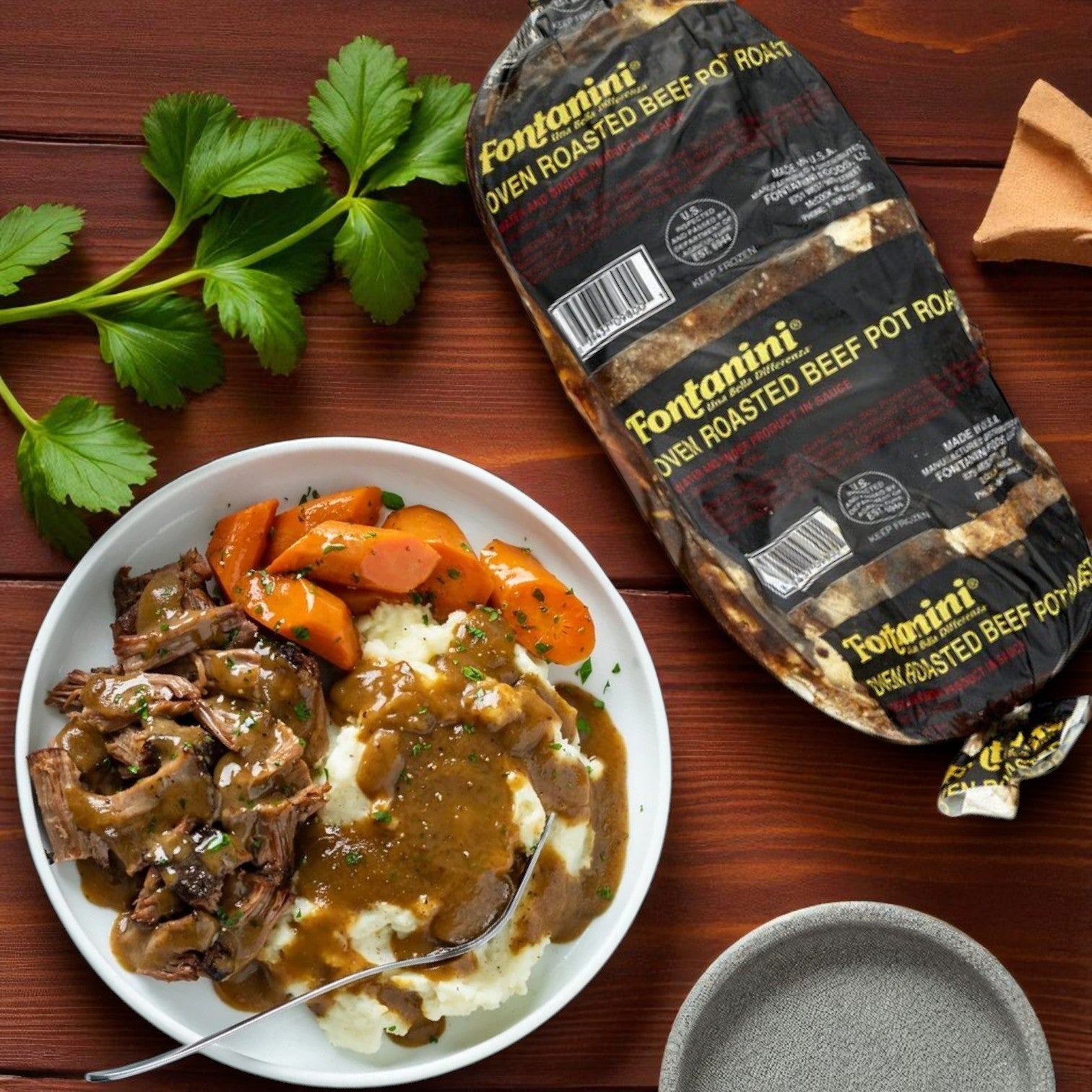 A plate with mashed potatoes, carrots, and beef pot roast with gravy sits beside Sysco’s Fontanini Beef Pot Roast with Gravy – 2 Packs (14–15 lbs Each), parsley, and an empty plate on a wooden table for homestyle comfort.