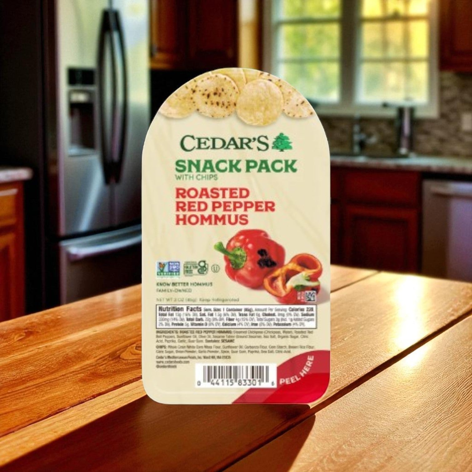 A Cedar's Roasted Red Peppers Hommus w/ Hommus Chips -1 Count sits on a kitchen table, offering a healthy snack option from Cedar's. A refrigerator and cabinets are visible in the background.