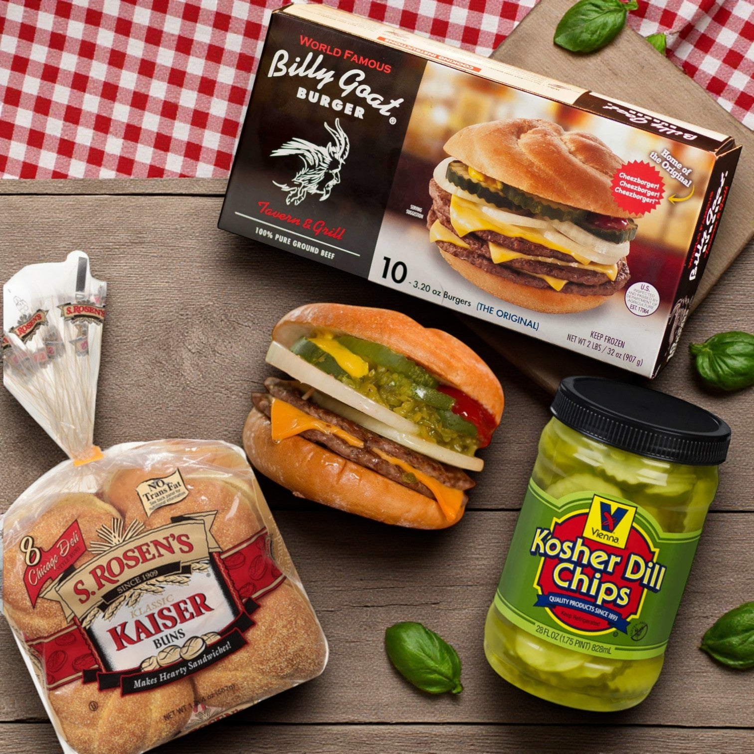 A box of Vienna Beef’s Billy Goat Tavern Burger Combo—featuring S. Rosen’s Kaiser Buns and Vienna Dill Chips—is placed on a wooden surface with basil leaves and a red checked cloth.