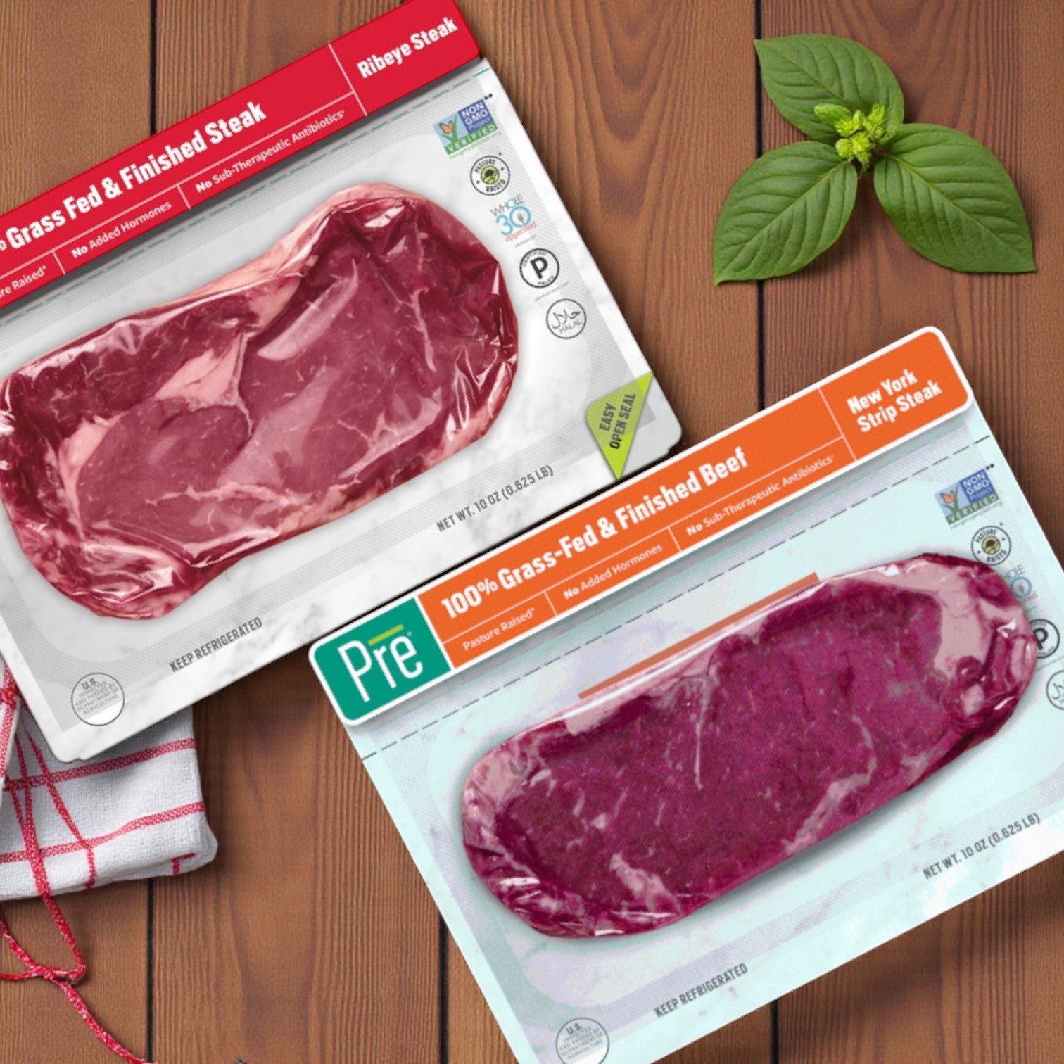 Two vacuum-sealed packs of Pre Brands 100% Grass Fed Steak Combo – Ribeye & New York Strip rest on a wooden surface with a basil sprig and red and white cloth nearby.