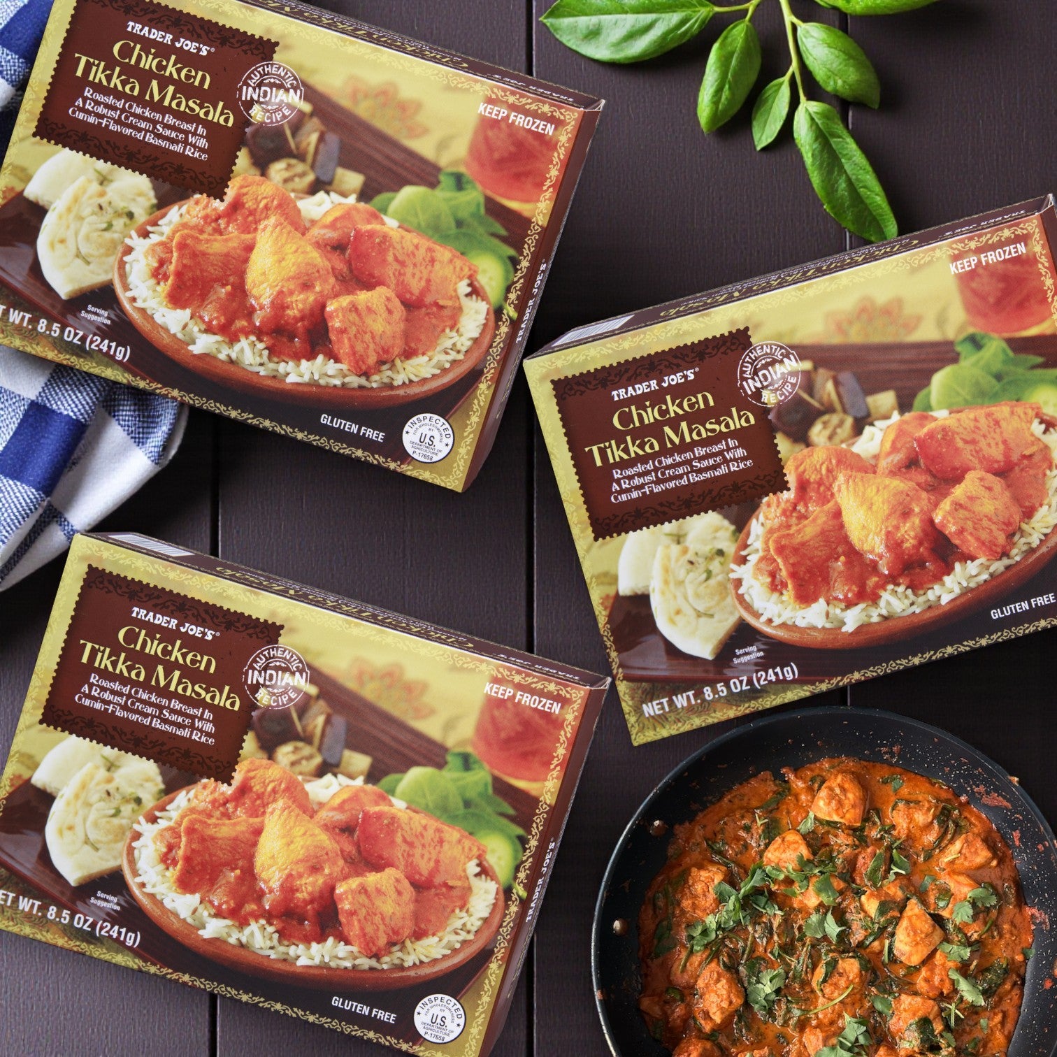 Three boxes of Easy Lunches Trader Joe’s Indian Cuisine Combo – 3 Pack Variety are displayed on a dark surface next to a dish of aromatic curry with basmati rice, finished with a blue cloth and green leaves for added color.