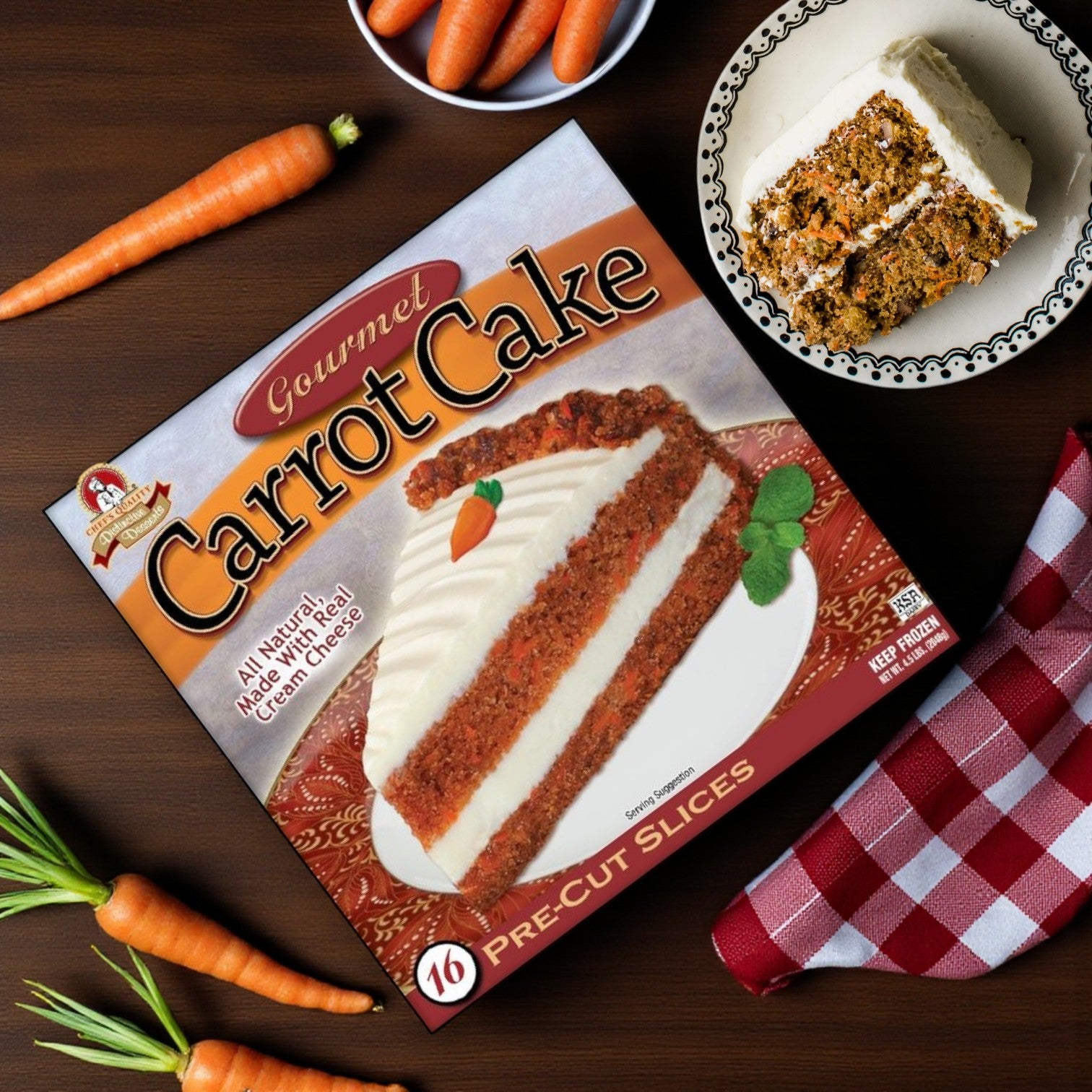 An Easy Lunches Frozen Chef’s Quality Carrot Cake with Cream Cheese Icing (16 slices, 4 lb) sits on a table beside a plate of cake, fresh carrots, and a red-checked napkin.