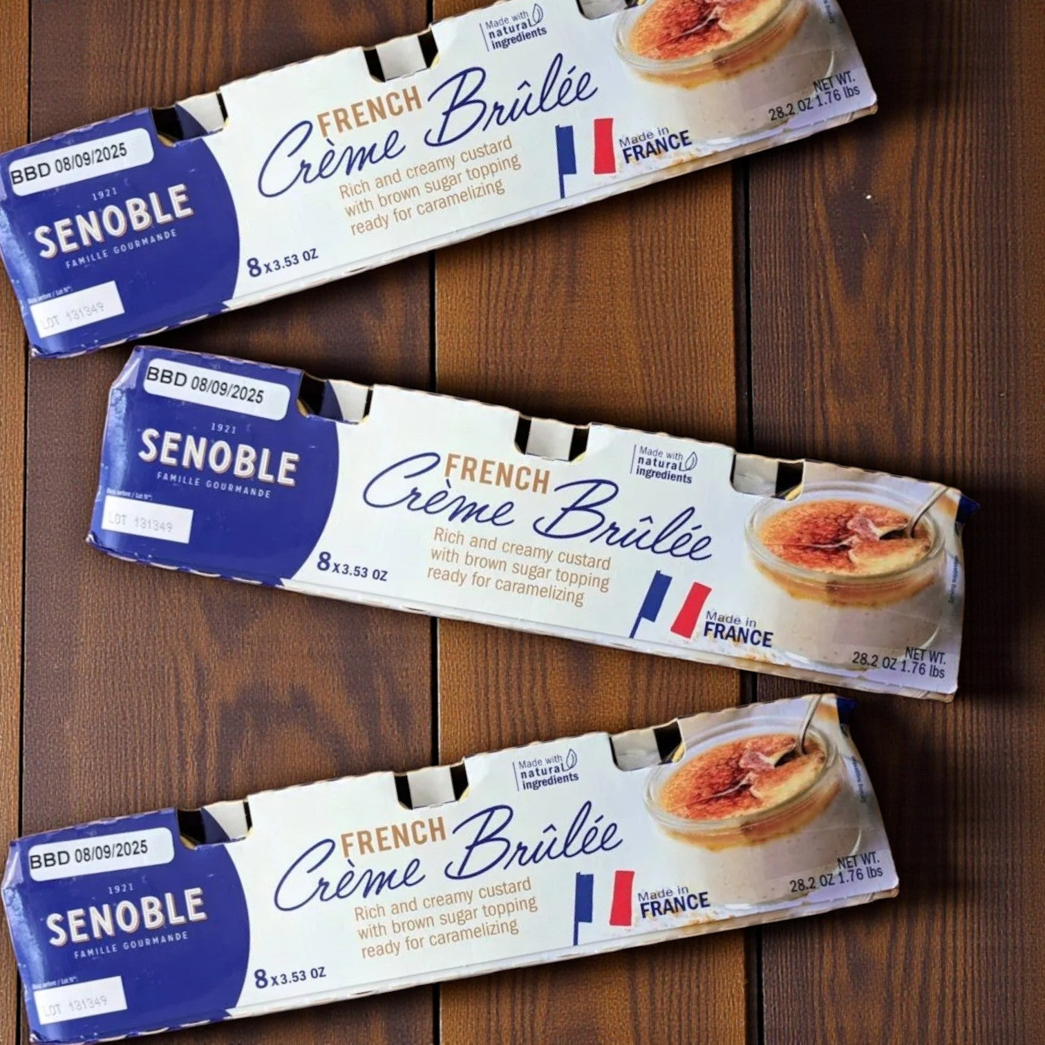 Three packs of Senoble French Crème Brûlée, each with eight 3.75 oz desserts, are displayed on a wooden surface, showcasing their creamy vanilla custard and caramelized sugar topping alongside the packaging.