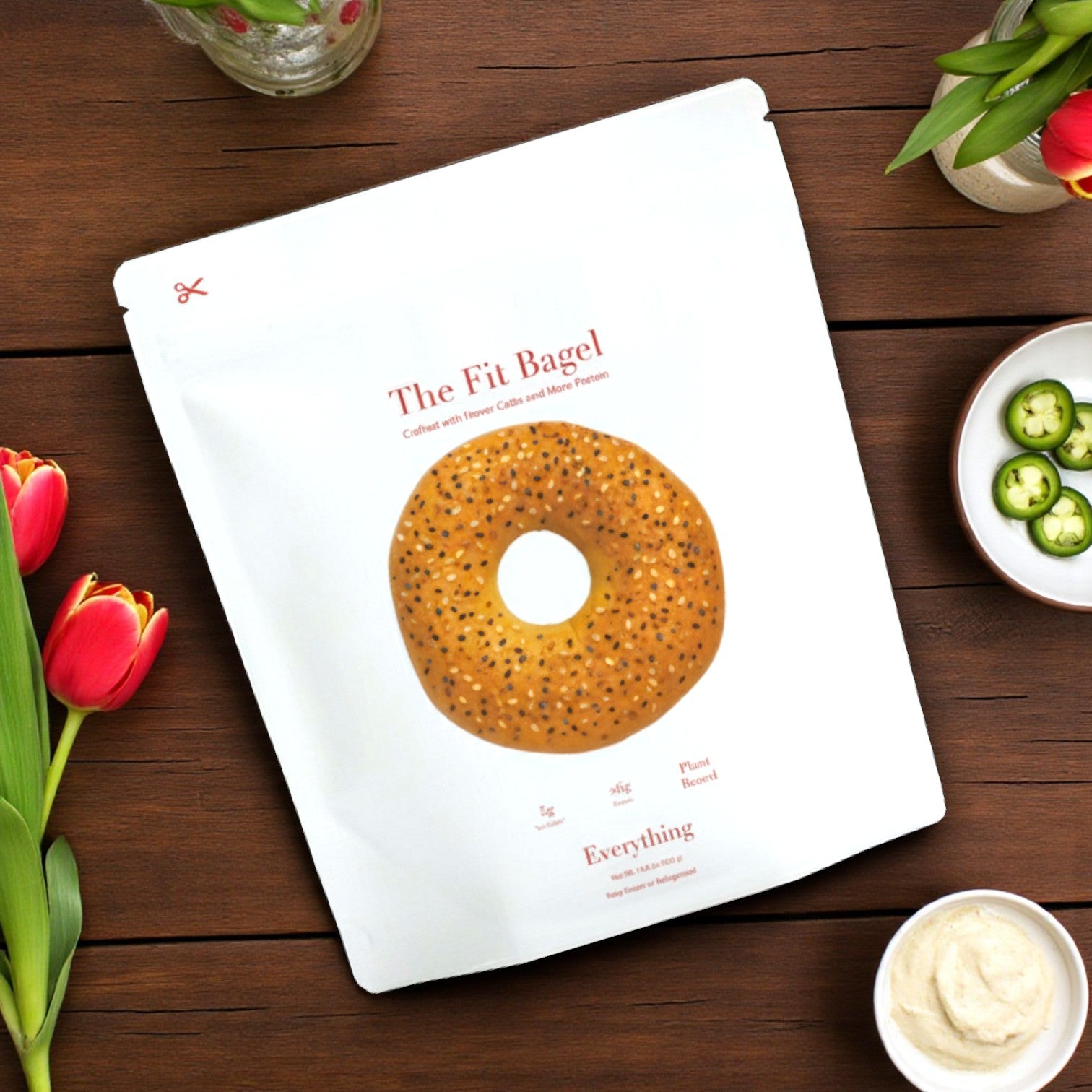 A package of The Fit Bagel’s Better Classic Bagel (4) & Fit Everything Bagel (4) Bundle (8 bags, 32 bagels) sits on a wooden table with flowers, cream cheese, and jalapeños, showcasing tasty, low net carb bagels perfect for a healthy snack.