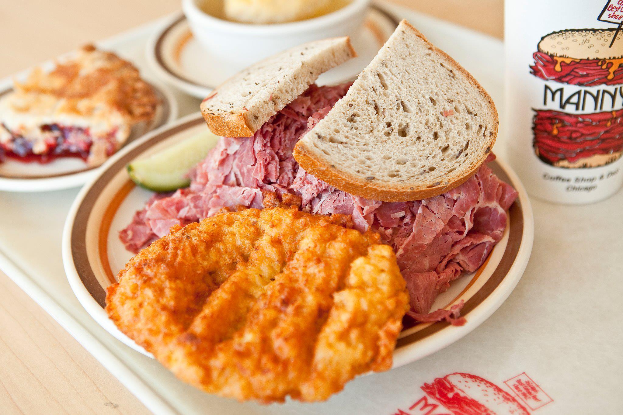 A plate with a pastrami sandwich, a hash brown, and a pickle slice, accompanied by a cherry turnover and a cup with "Manny's" branding.